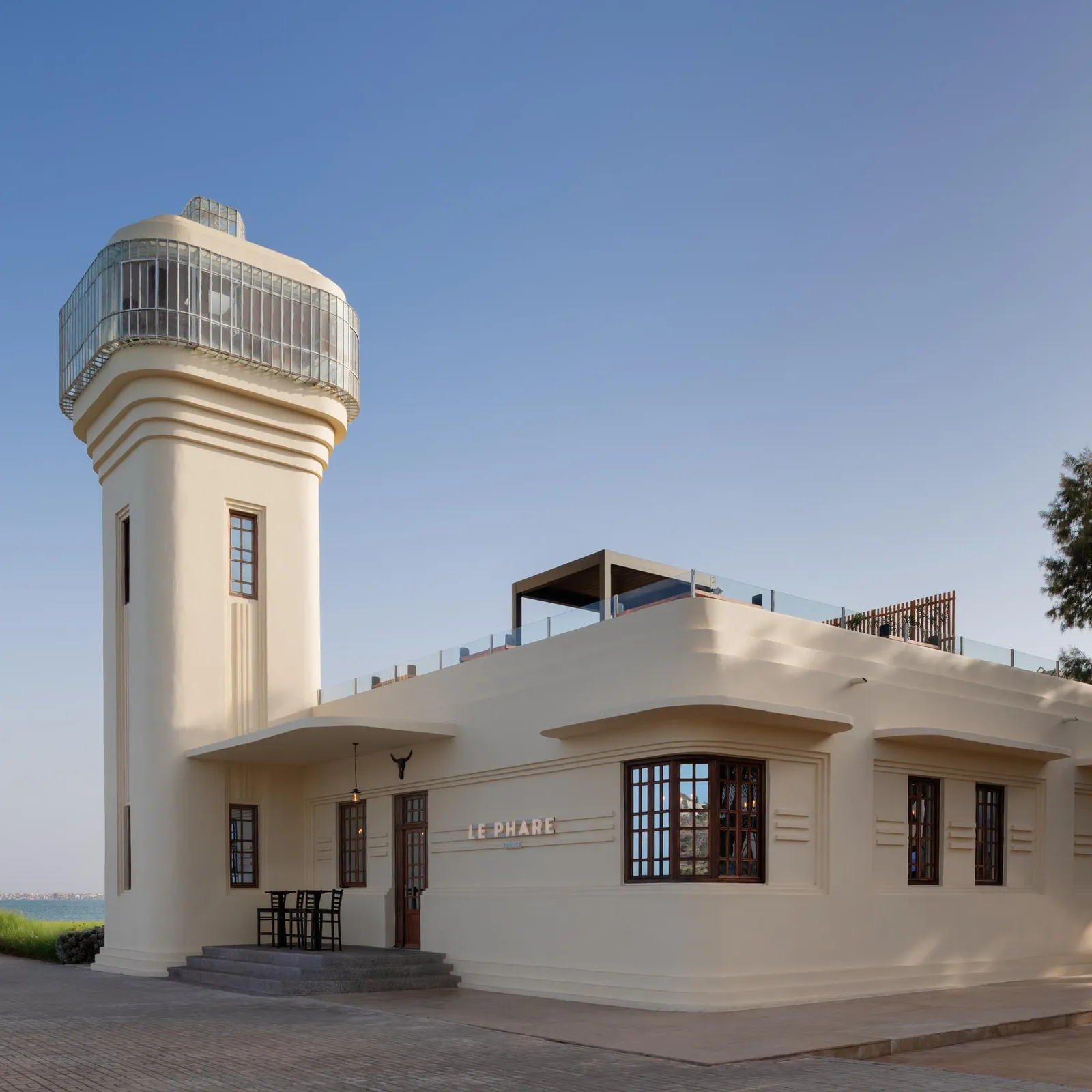 A white lighthouse with a square tower and a glass observation platform, located near the sea, with a low adjacent building bearing the sign "LE PHARE"