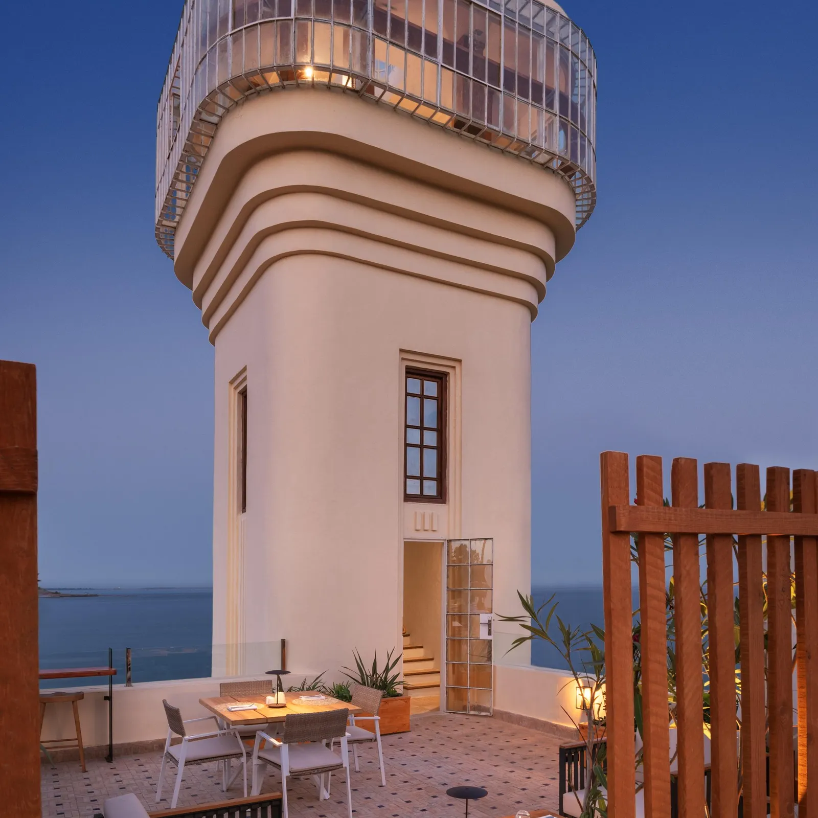 A white building with a glass-domed terrace overlooks the sea, with tables and chairs arranged on a stone terrace.