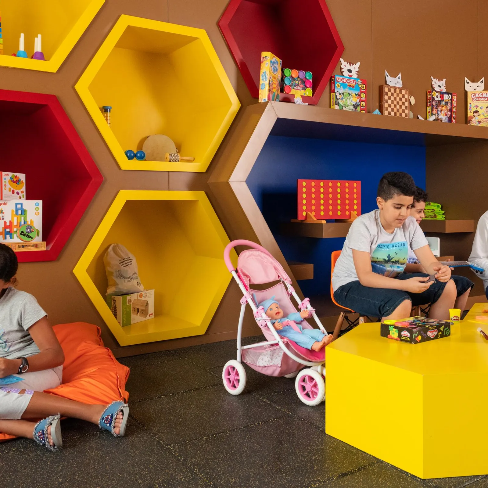 Children reading and playing in a colorful room with hexagonal shelves filled with toys and books.