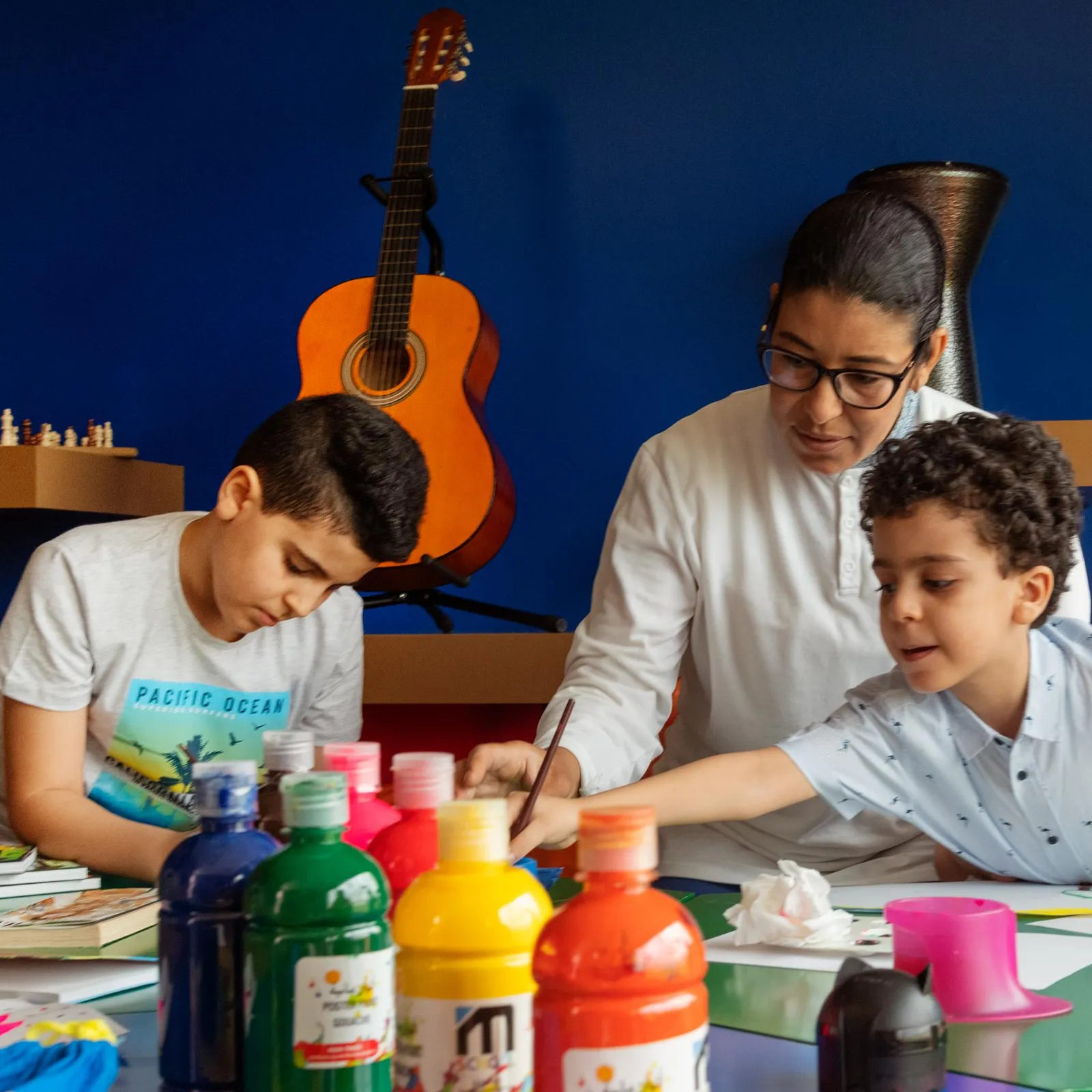 Three people are sitting at a table and painting with colored paint bottles; a guitarist is visible in the background.