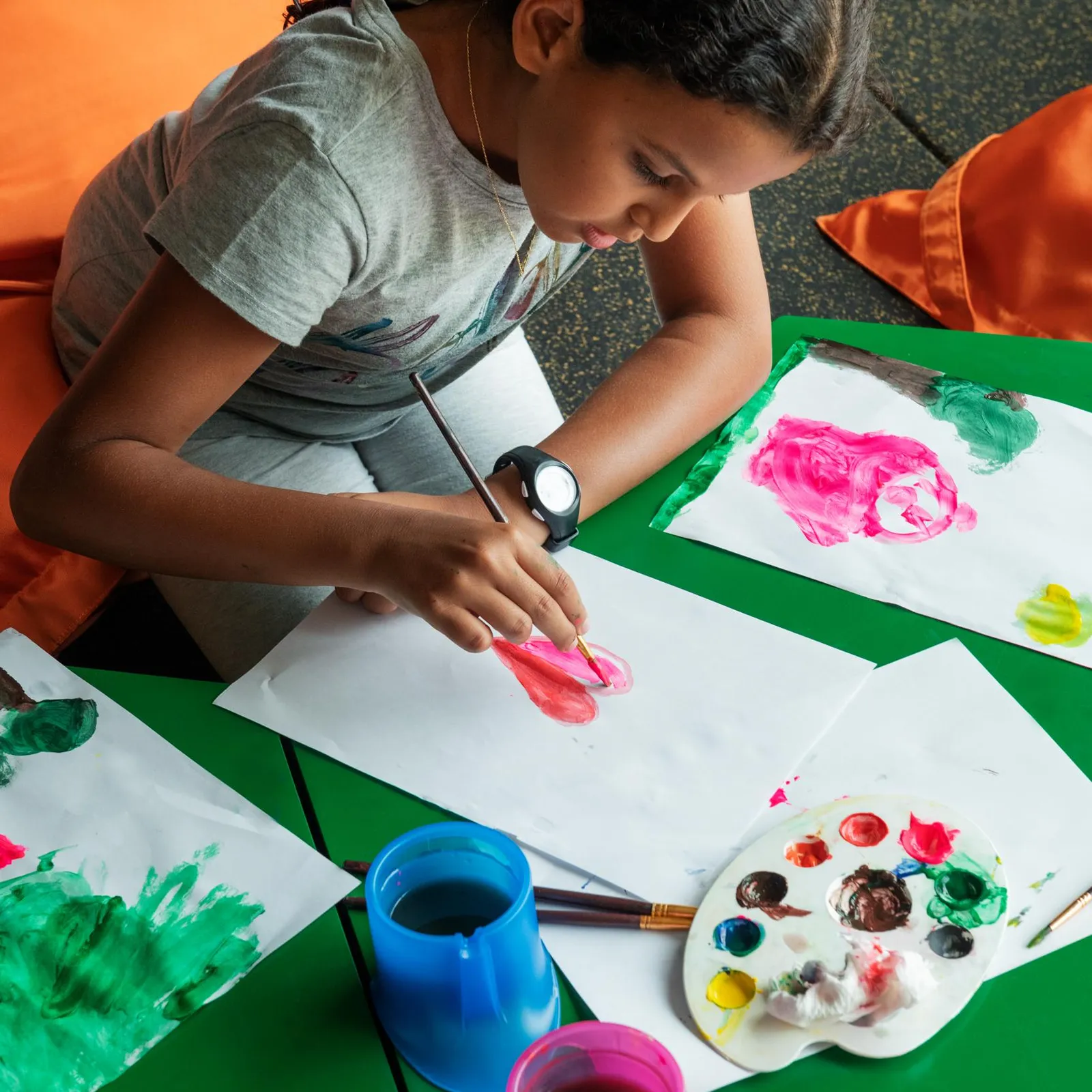 Child painting on sheets of paper with brushes and paint on a green table