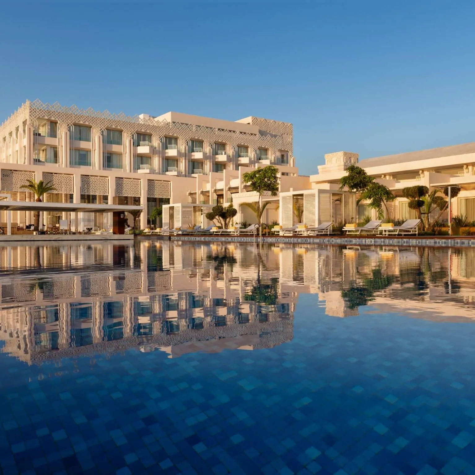 A large white building with balconies and geometric patterns, reflected in a blue pool, surrounded by palm trees and lounge chairs.