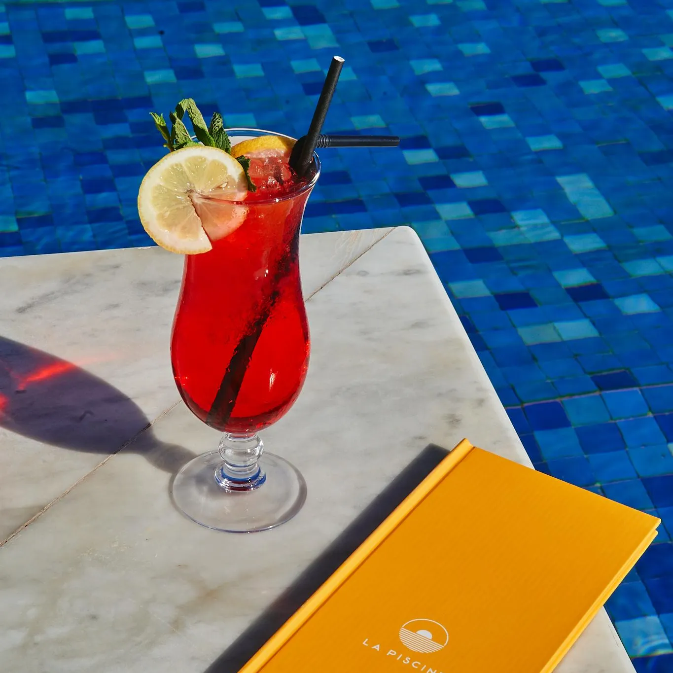 A glass of red drink with a lemon slice and mint leaves placed on a table near a pool, next to a yellow menu