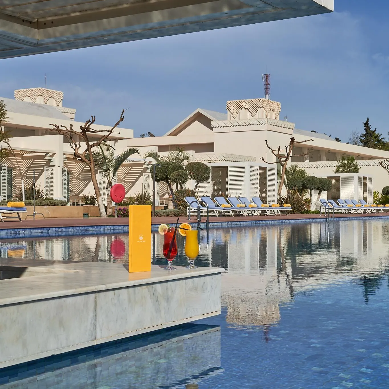 Pool with two glasses of colorful drinks on the edge, surrounded by sun loungers and white buildings with flat roofs
