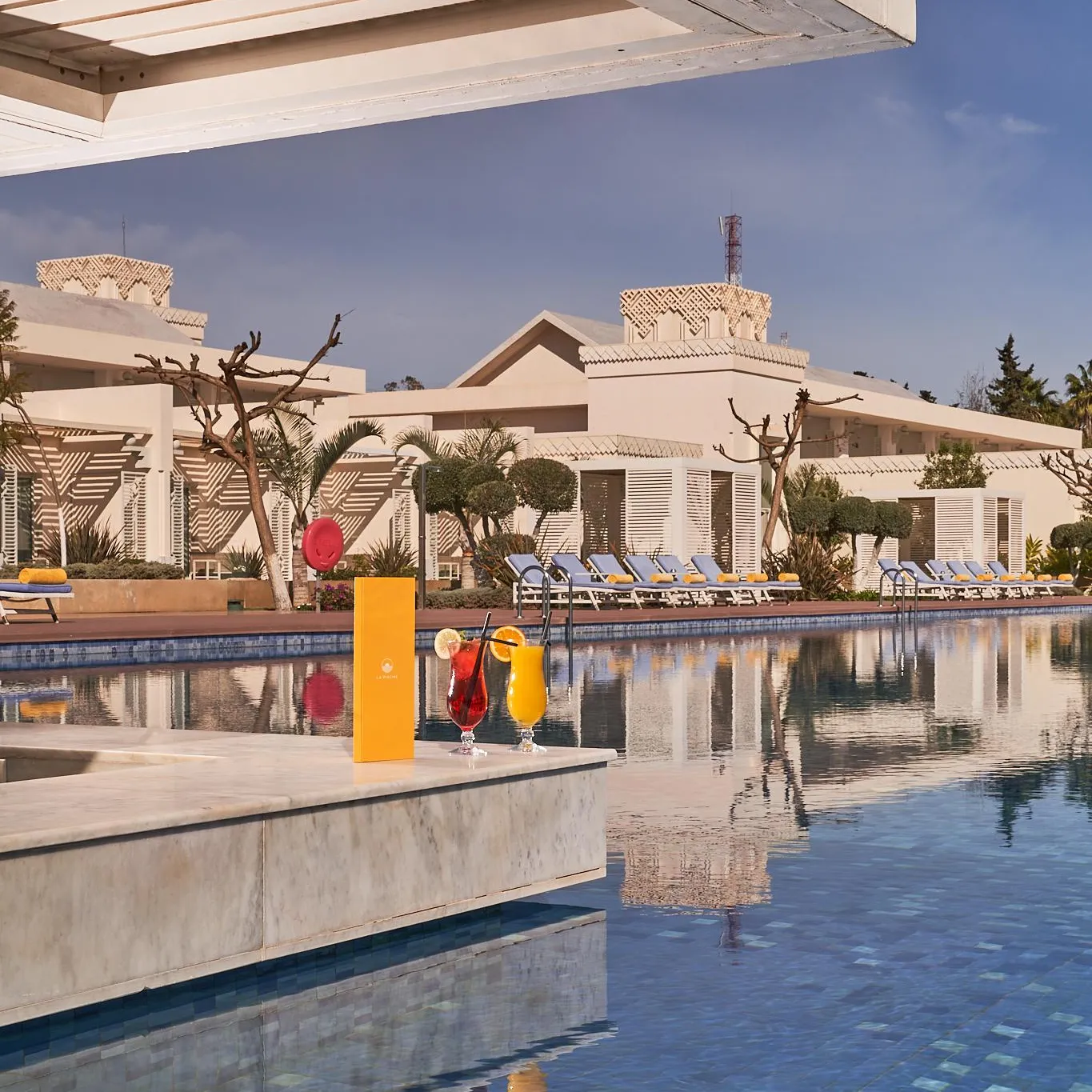 Two glasses of colorful drinks on a table near a pool with a beige building in the background.