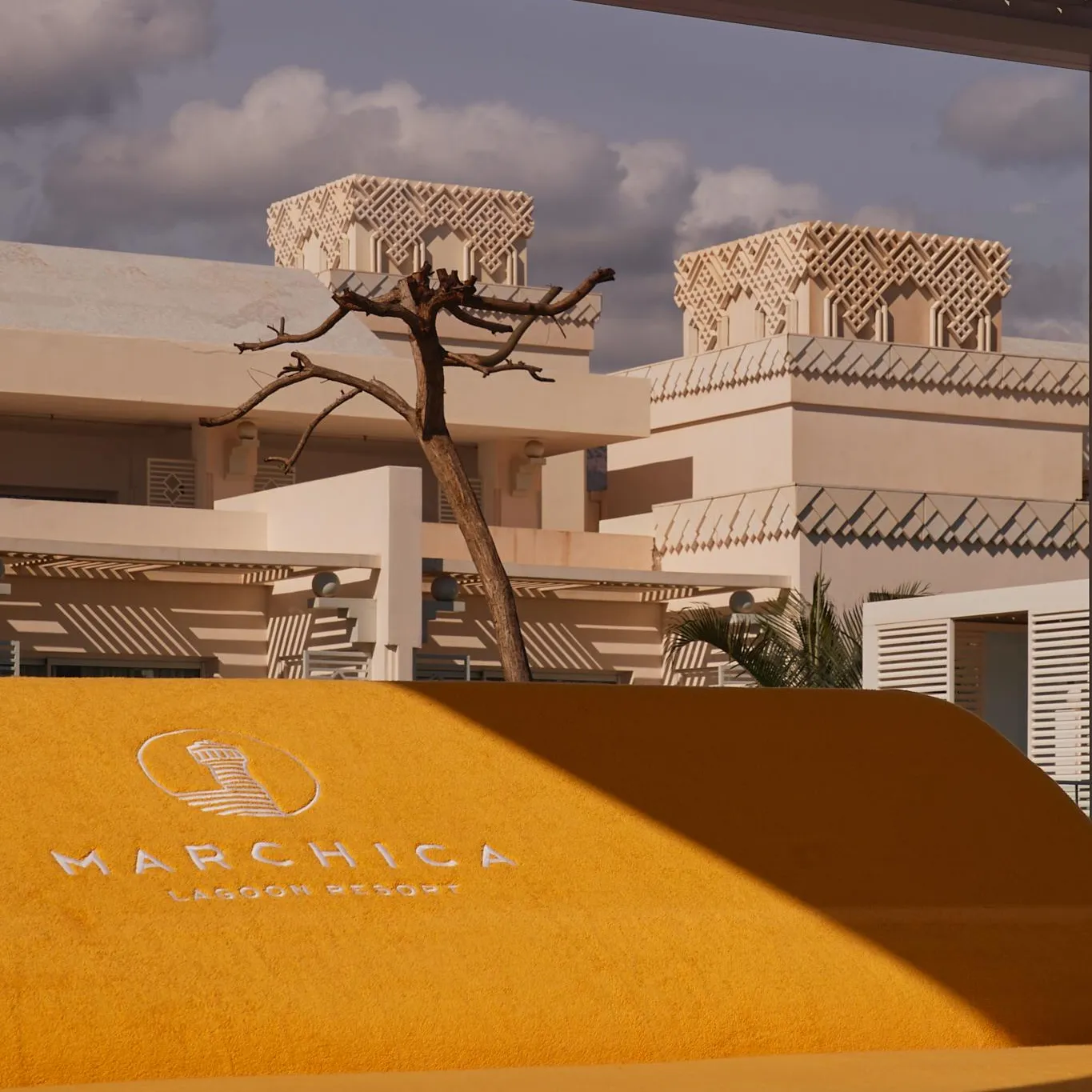 A beige building with balconies and geometric patterns on the roofs, under a partly cloudy sky, with a "Marchica Lagoon Resort" sign in the foreground.