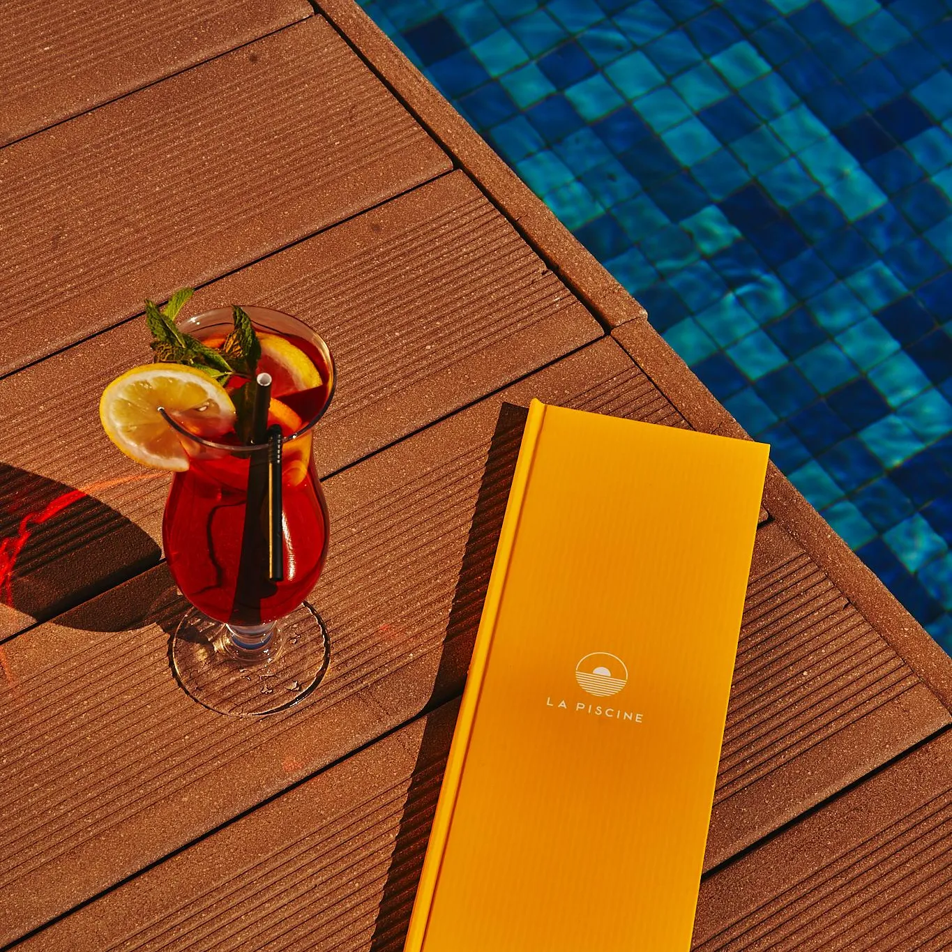 A glass of red drink with an orange slice and a straw, a yellow menu, and a blue pool in the background.