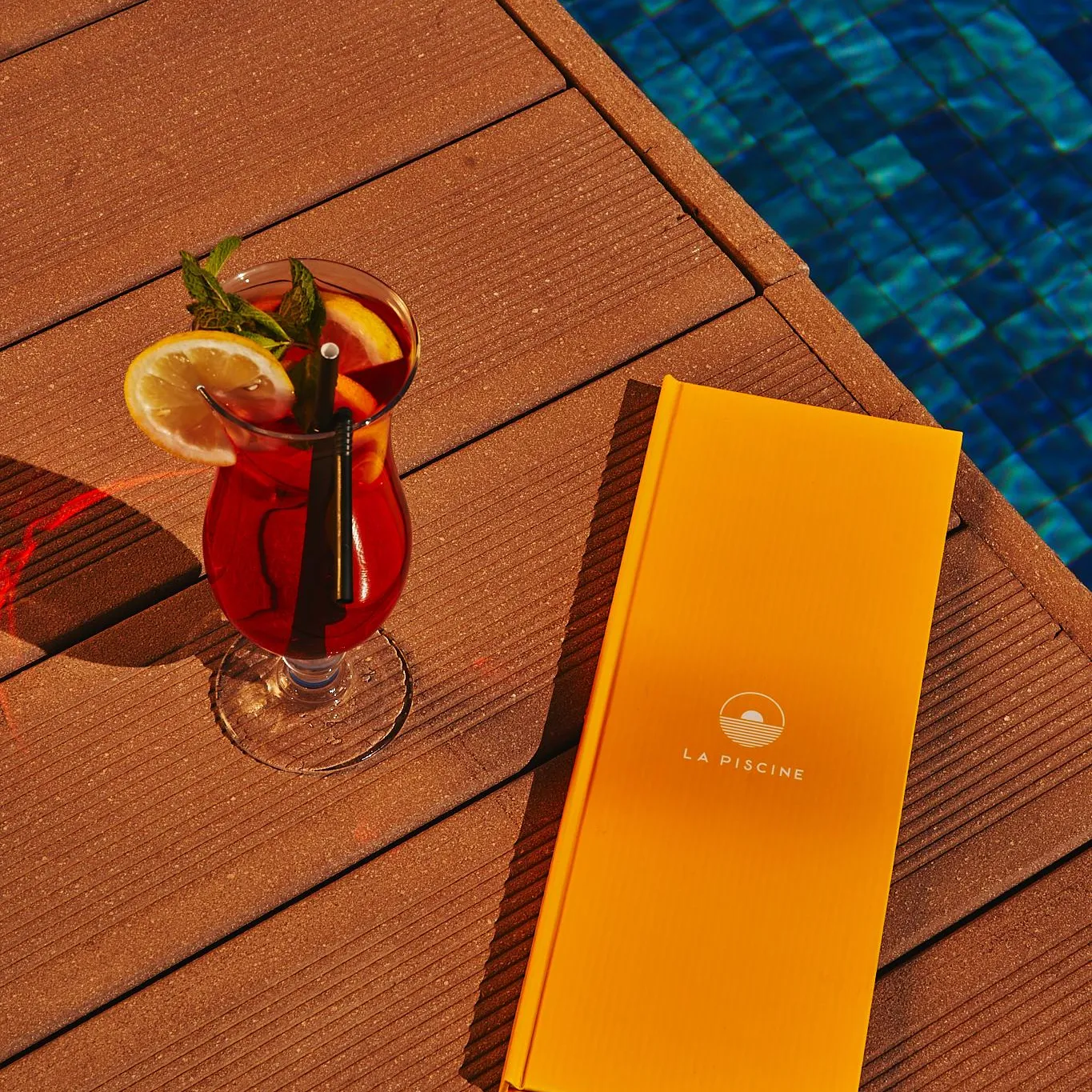 A glass of red drink with an orange slice and a straw, an orange menu, and a blue pool in the background.
