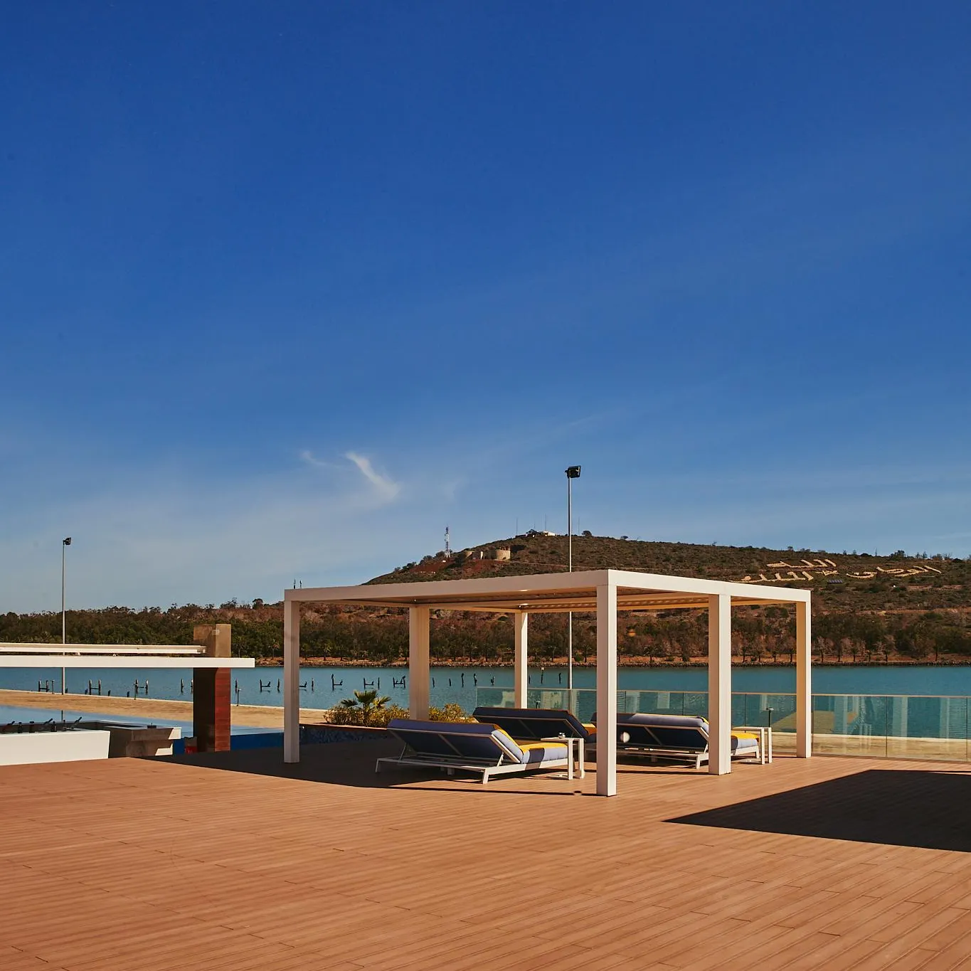 White pergola with sun loungers on a wooden terrace near a swimming pool and a body of water with hills in the background