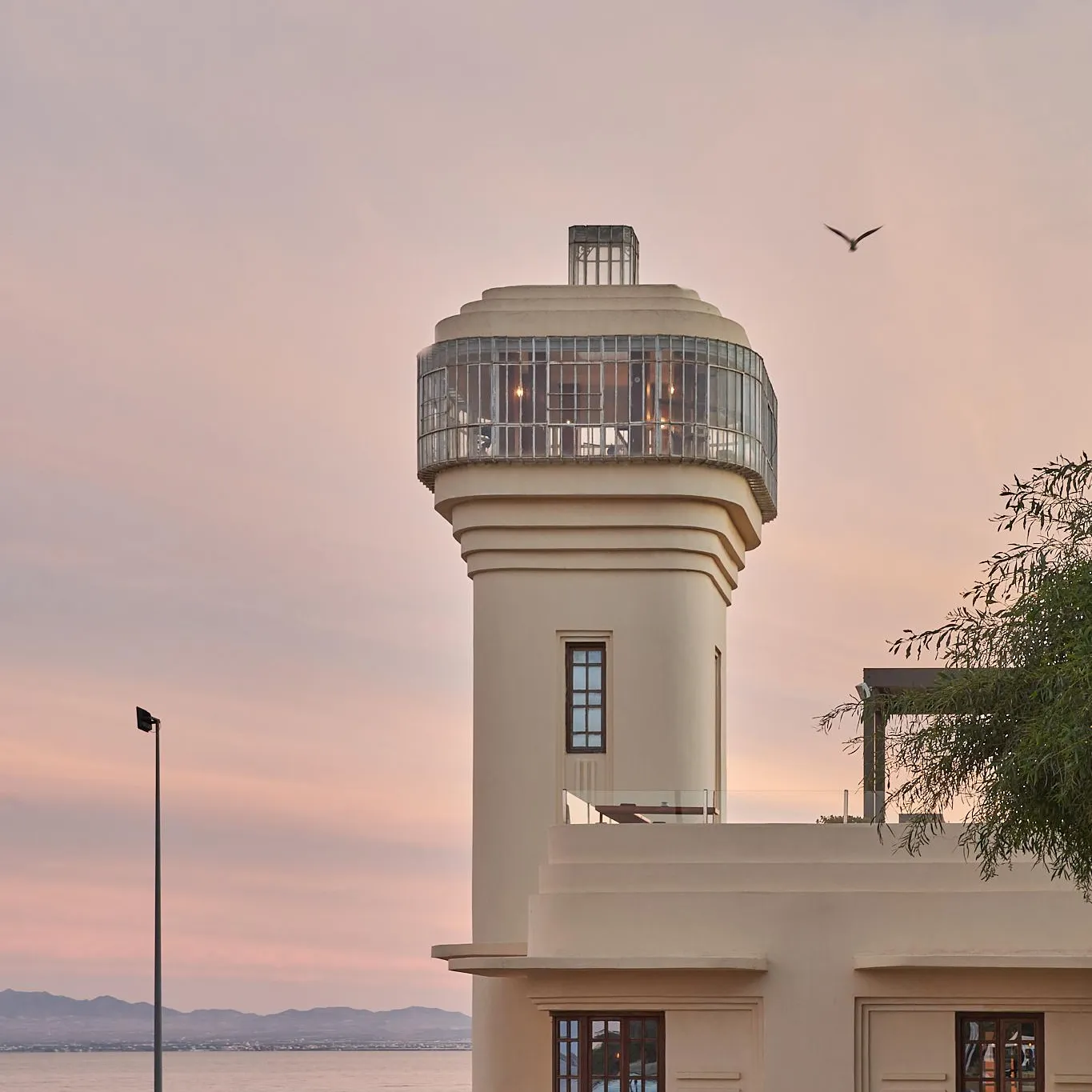 A modern lighthouse with a glass lantern at the top stands near a body of water at sunset.