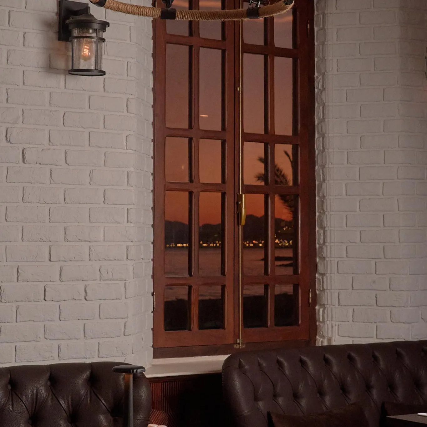 A table with cutlery and candles, under a cord and bulb light fixture, in front of a wooden-framed window and a white brick wall.