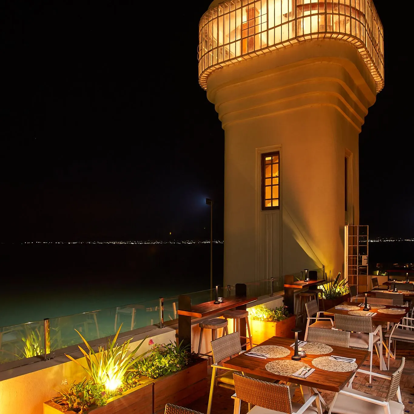 Restaurant terrace lit at night with tables, chairs, and plants, located near a lit cylindrical structure and a pool.