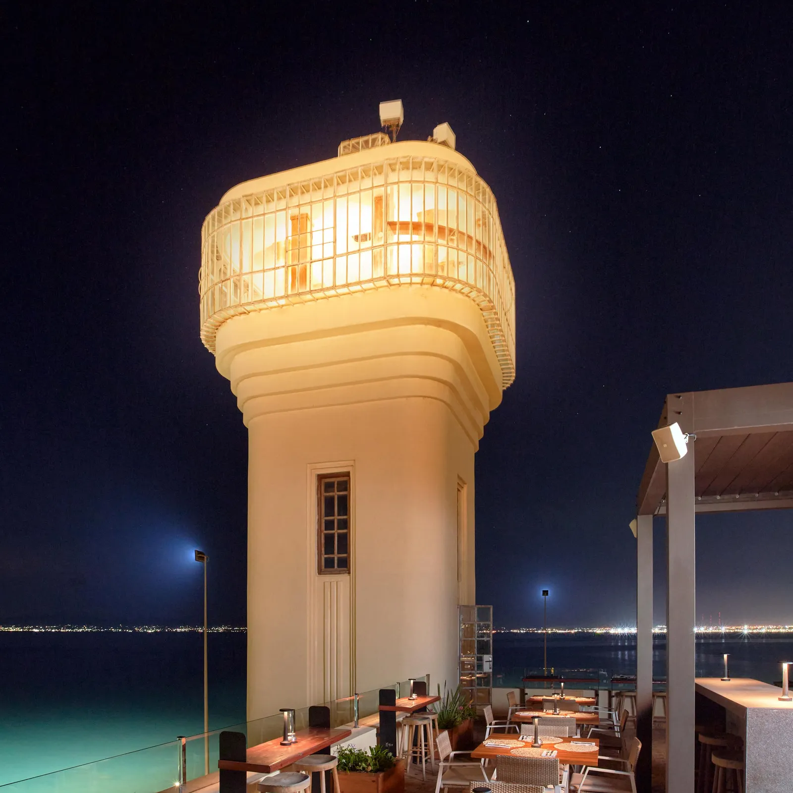 A seaside restaurant terrace with tables and chairs, lit at night, located near a tower with an illuminated lattice structure.
