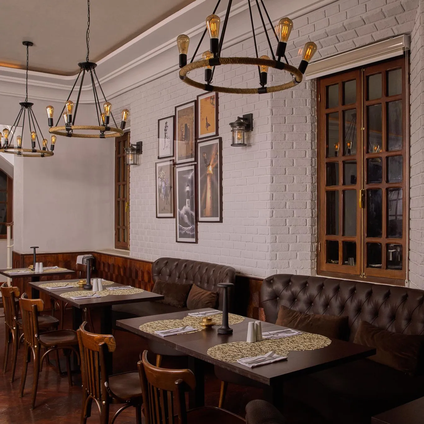 Restaurant with wooden tables, leather chairs and banquettes, lit by pendant lamps with bare bulbs, white brick walls and wooden window frames.