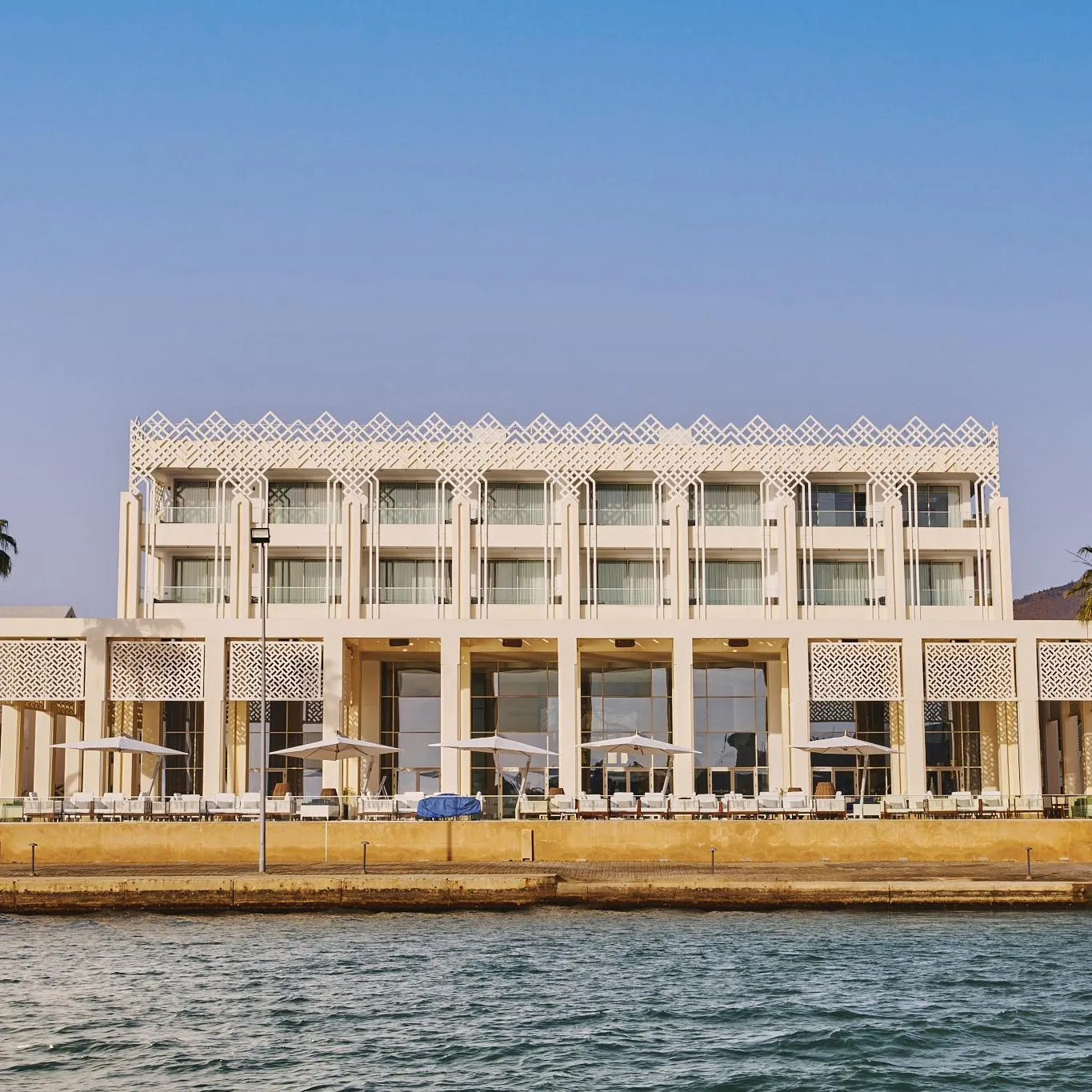 A white modern-style building with balconies and columns, located by the water with palm trees and tables under umbrellas.