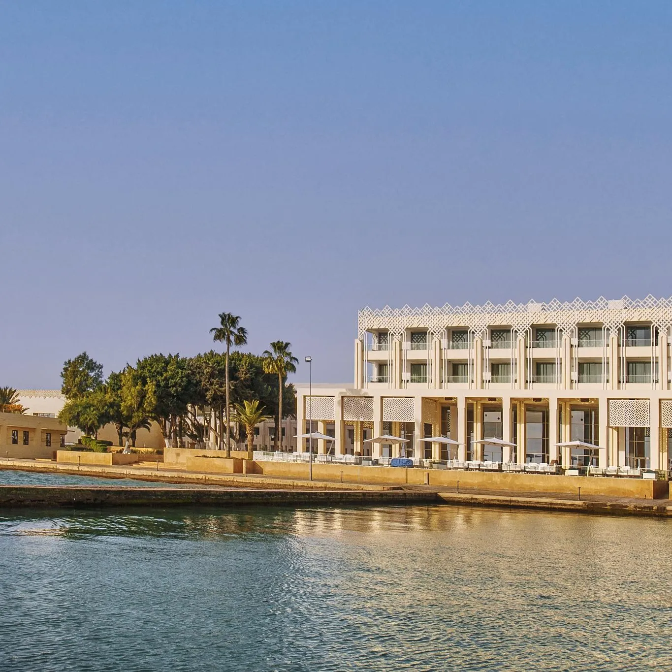 A modern building with arches and balconies overlooks a calm expanse of water, surrounded by palm trees and other trees.