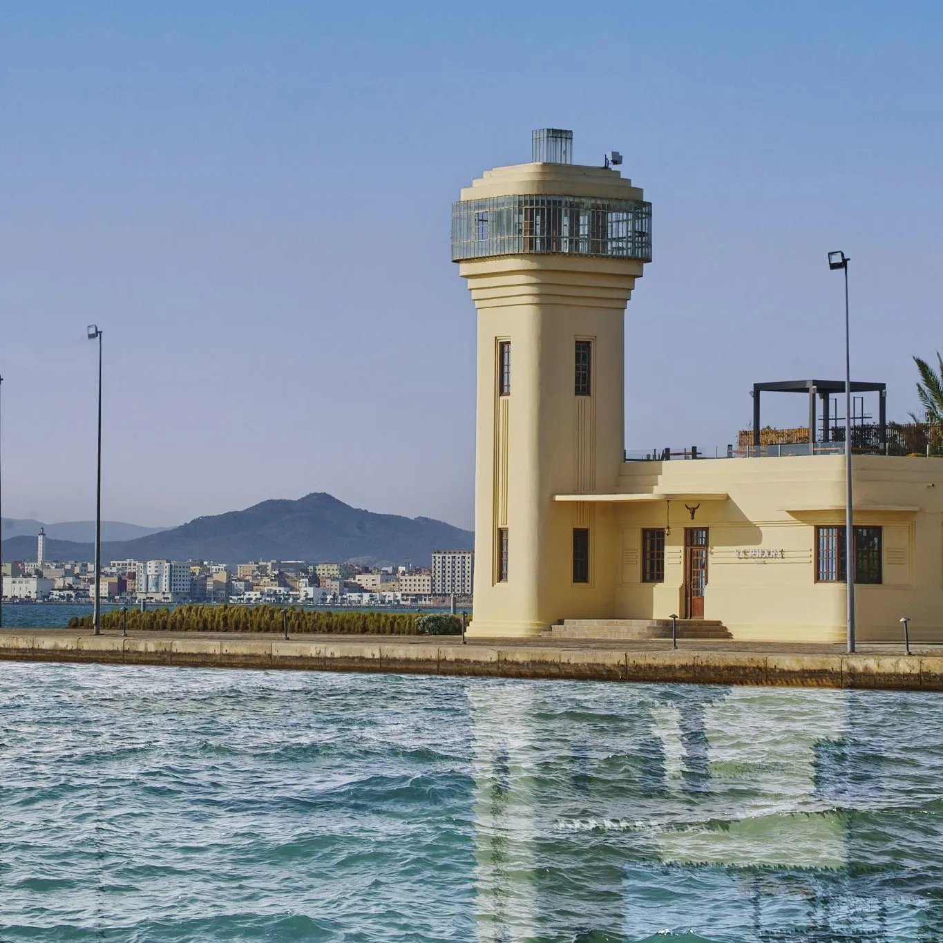Yellow control tower with an observatory on top, located by the water with buildings and mountains in the background