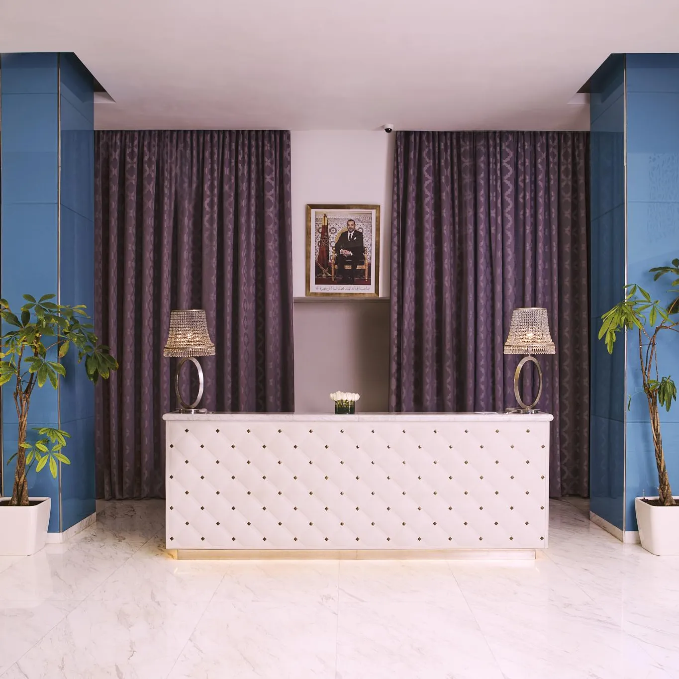 Hotel reception with a white counter, two lamps, and three green plants in front of purple curtains