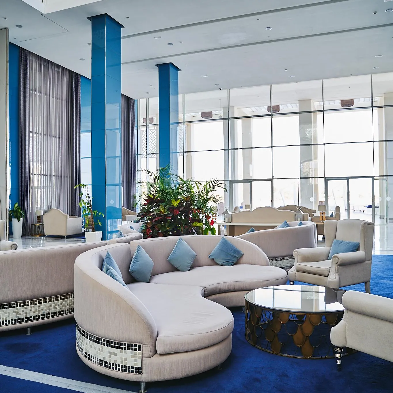 A modern hotel lounge with beige sofas and armchairs, blue cushions, a coffee table, and green plants, in front of large windows.