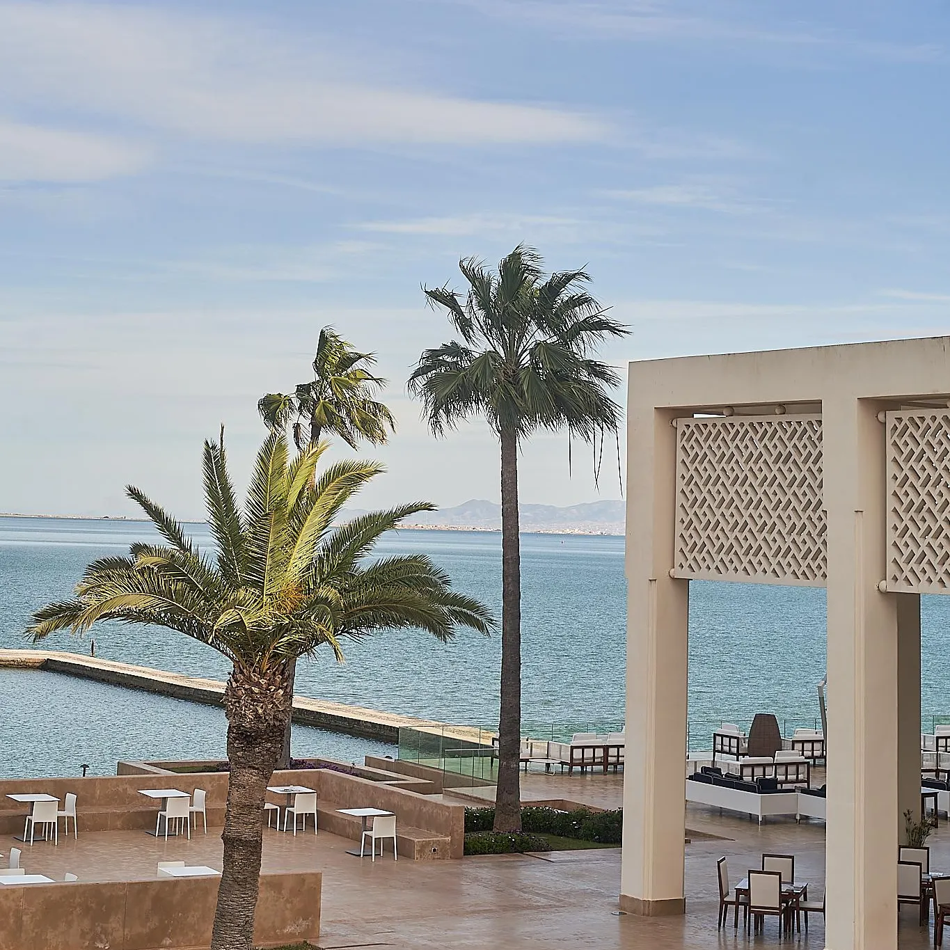 Terrace with tables and chairs near the sea, bordered by palm trees and an architectural structure with geometric patterns.