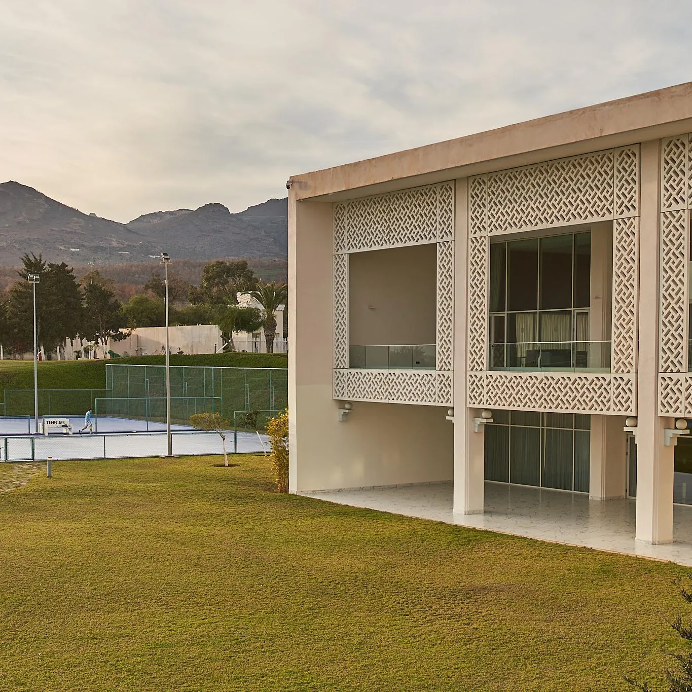 Modern building with geometric-patterned facade, surrounded by a lawn and tennis courts, with mountains in the background.