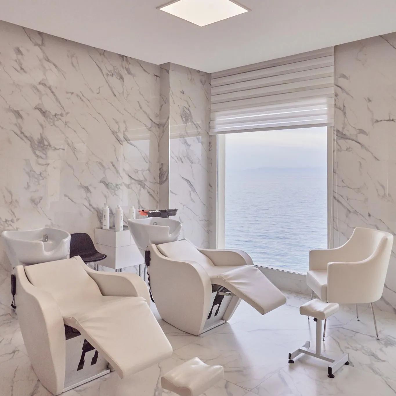 Hair salon with white hairdressing chairs and marble-covered white walls, offering a sea view through a large window.