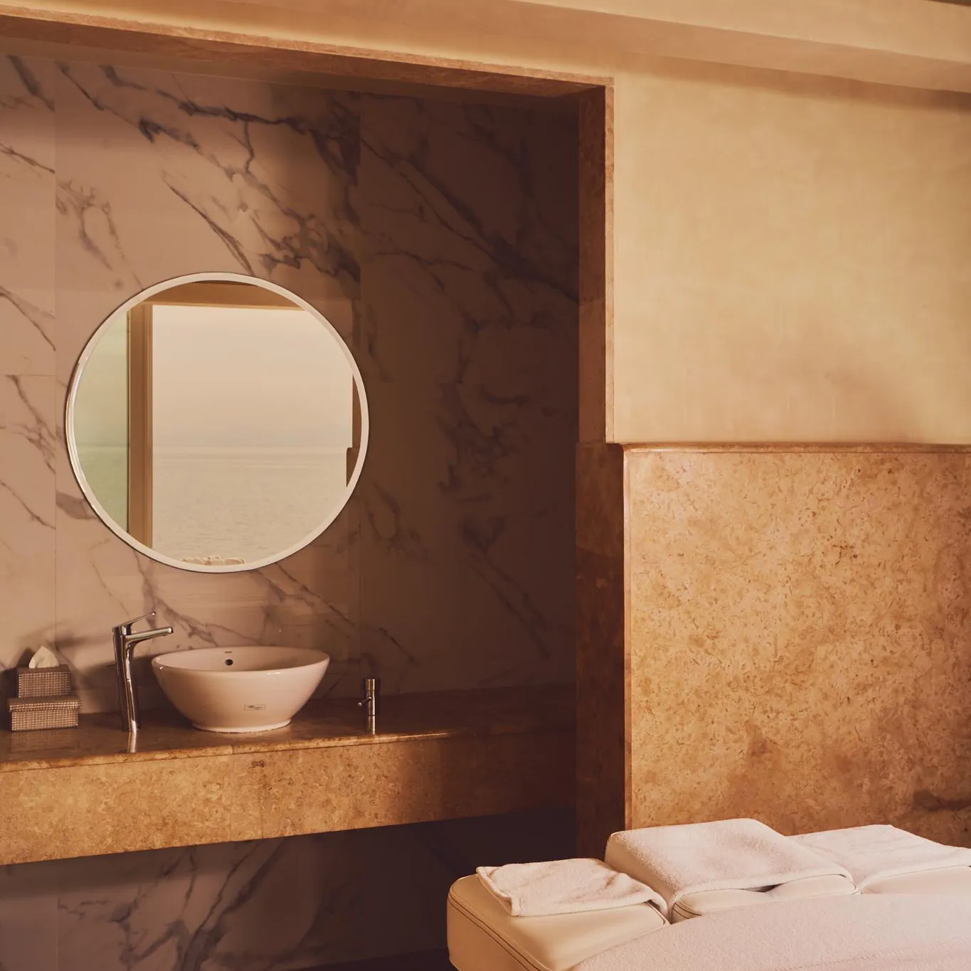 A sink built into a stone countertop with a round mirror above it, next to a massage table covered with white towels.