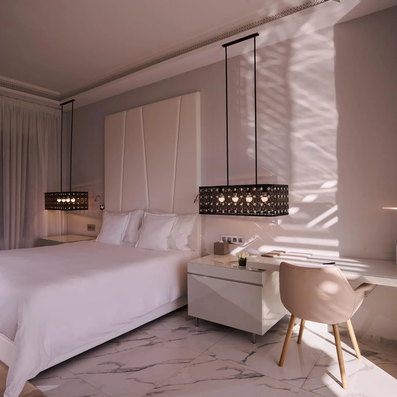 Room with a white double bed, a bedside table, and a chair near a window with translucent curtains.