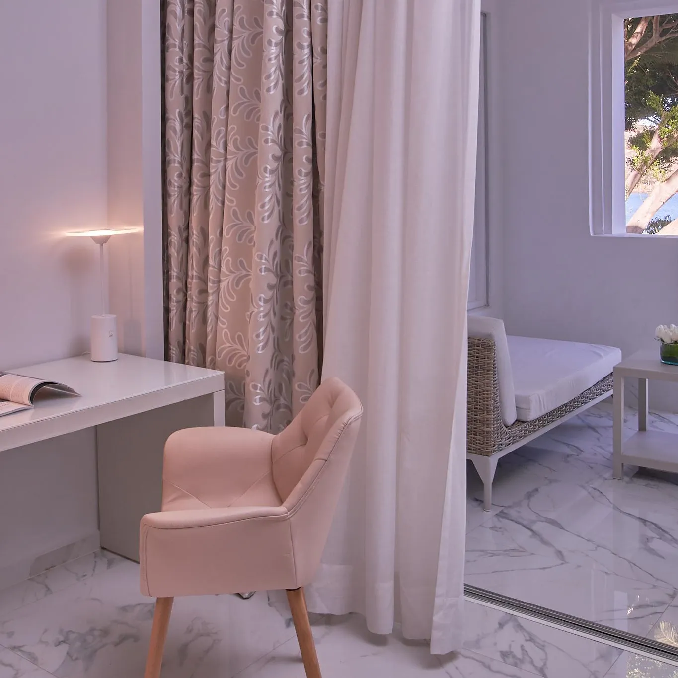 White desk with pink chair, lit lamp, magazines, and floral decoration near a window overlooking a garden with trees.