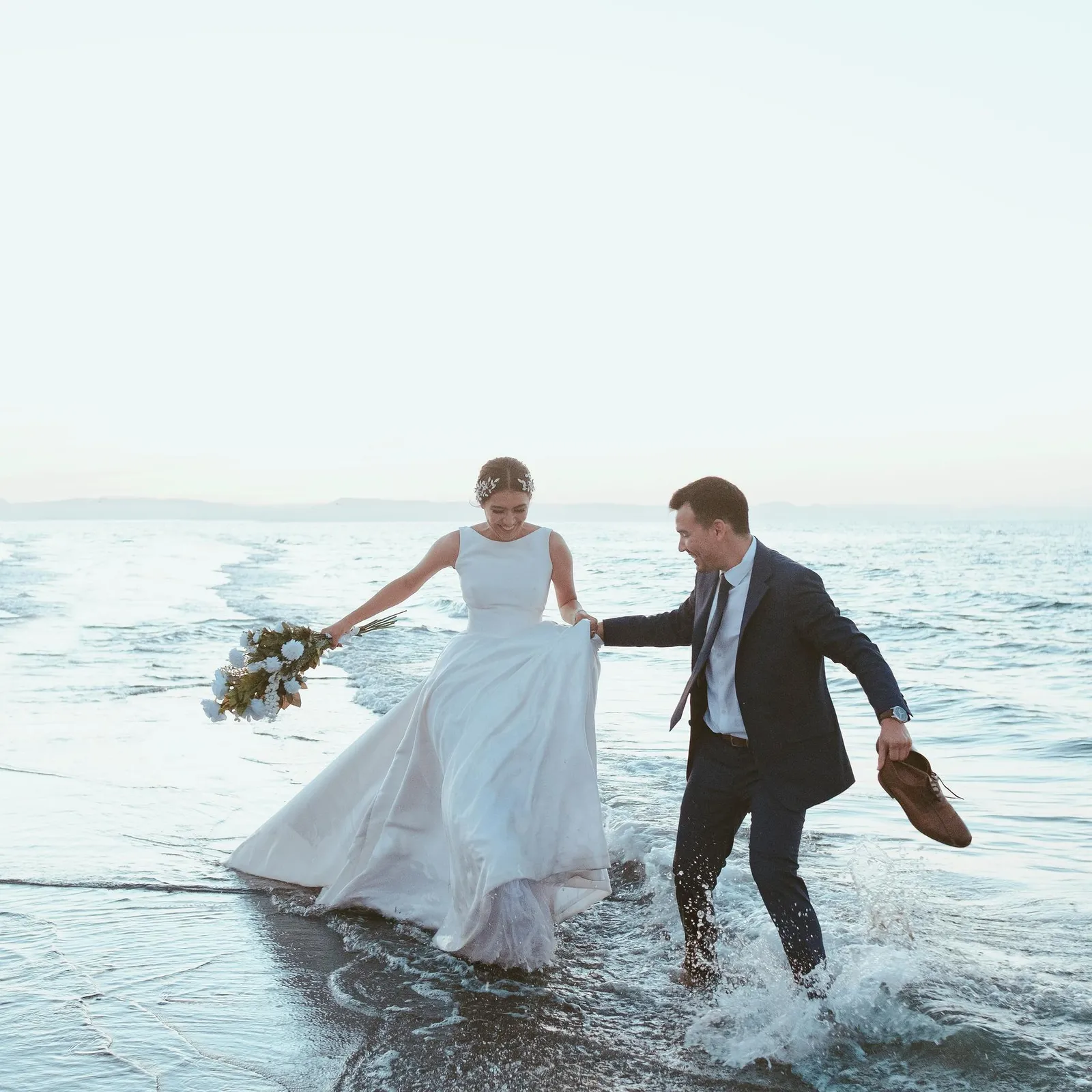 A couple walks barefoot in the water, the bride holding a bouquet and the man holding his shoes.