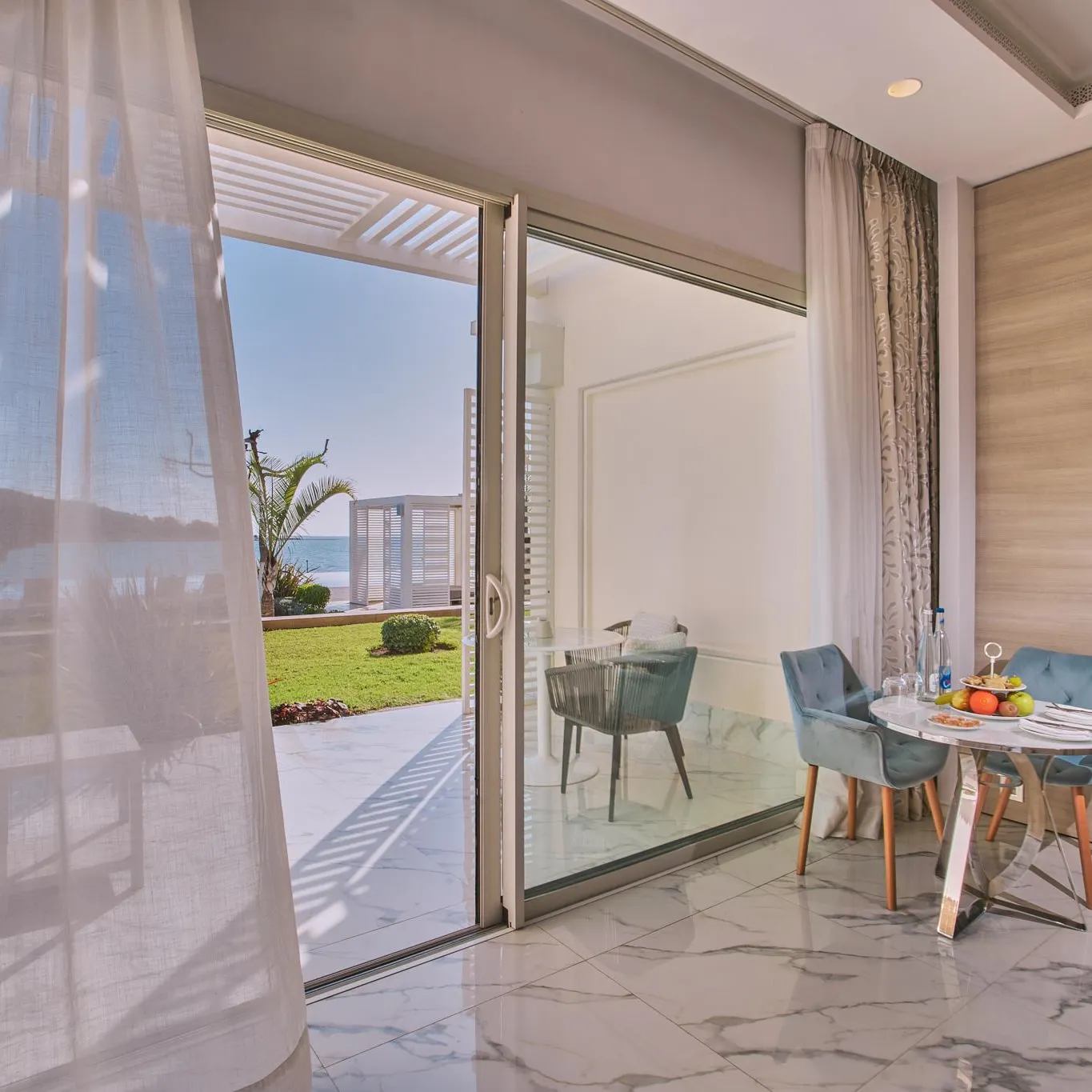A table with fruits and glasses is set up near a glass door open to a balcony with a sea view.