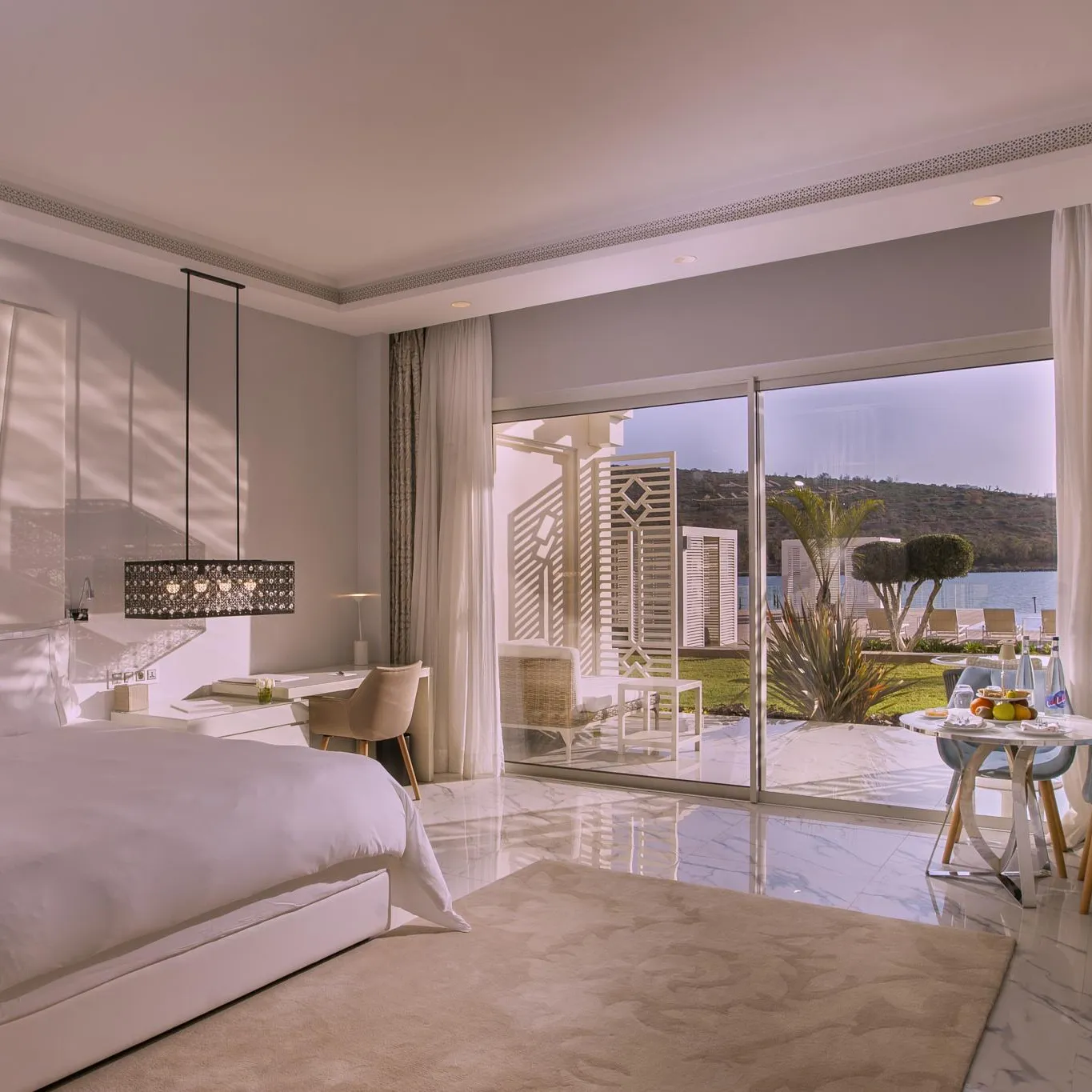 Luxurious room with white bed, work desk, and view of a terrace and outdoor landscape through large windows.