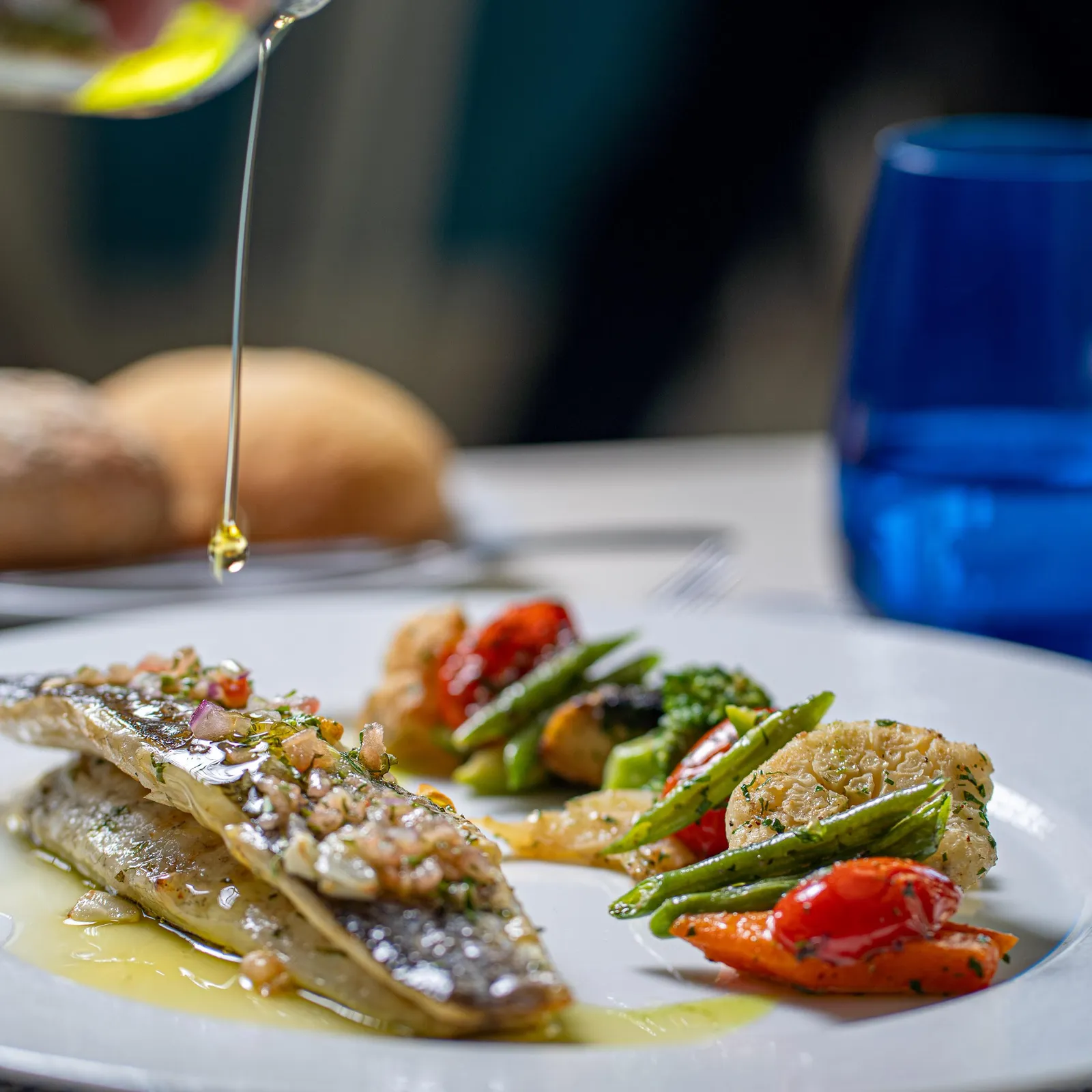 A hand pours oil over a dish consisting of grilled fish and vegetables, accompanied by bread.