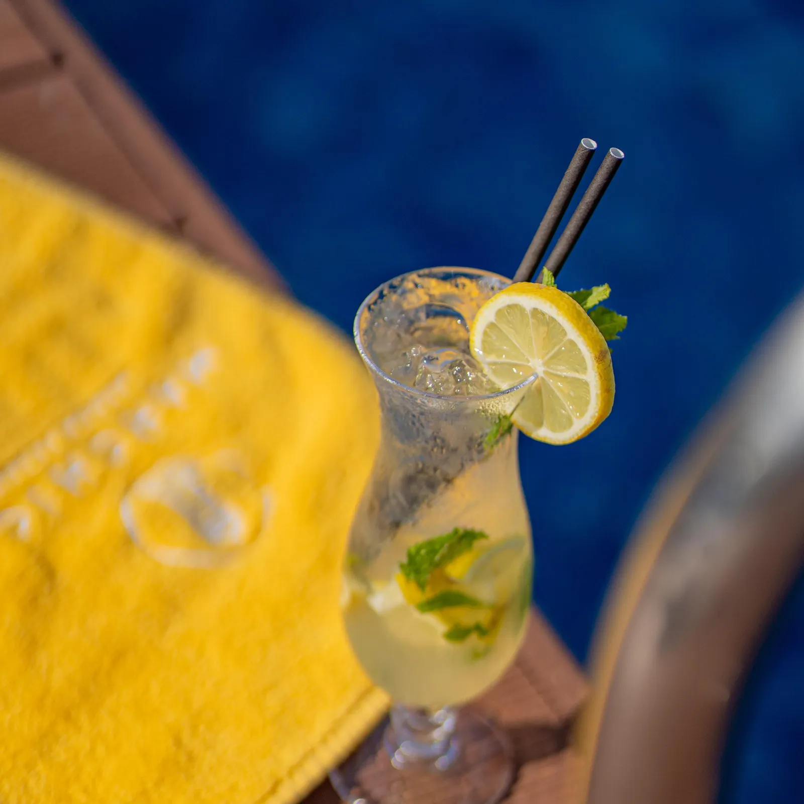 A glass of cold drink with a lemon slice and mint leaves, placed on a wooden table next to a yellow napkin.