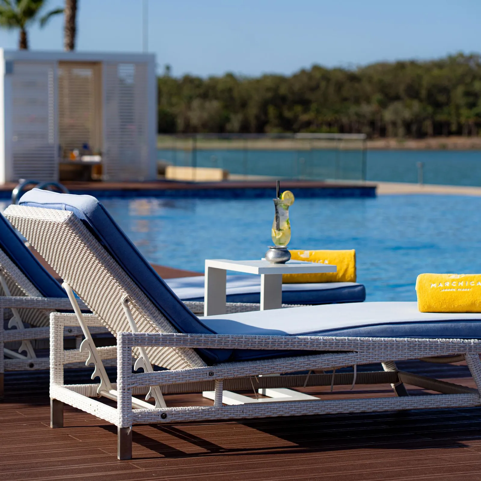 Two wicker lounge chairs with blue cushions and a yellow towel are arranged near a pool, along with a coffee table and a glass containing a drink.