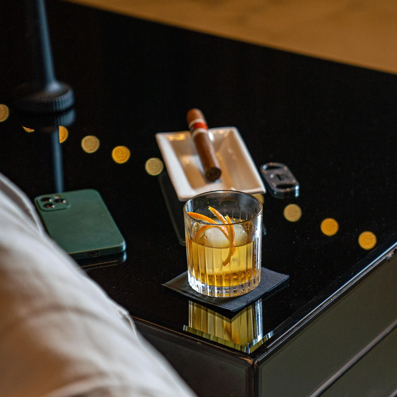 A glass of cocktail with ice cubes and an orange slice on a table, next to a smartphone and a cigar case.