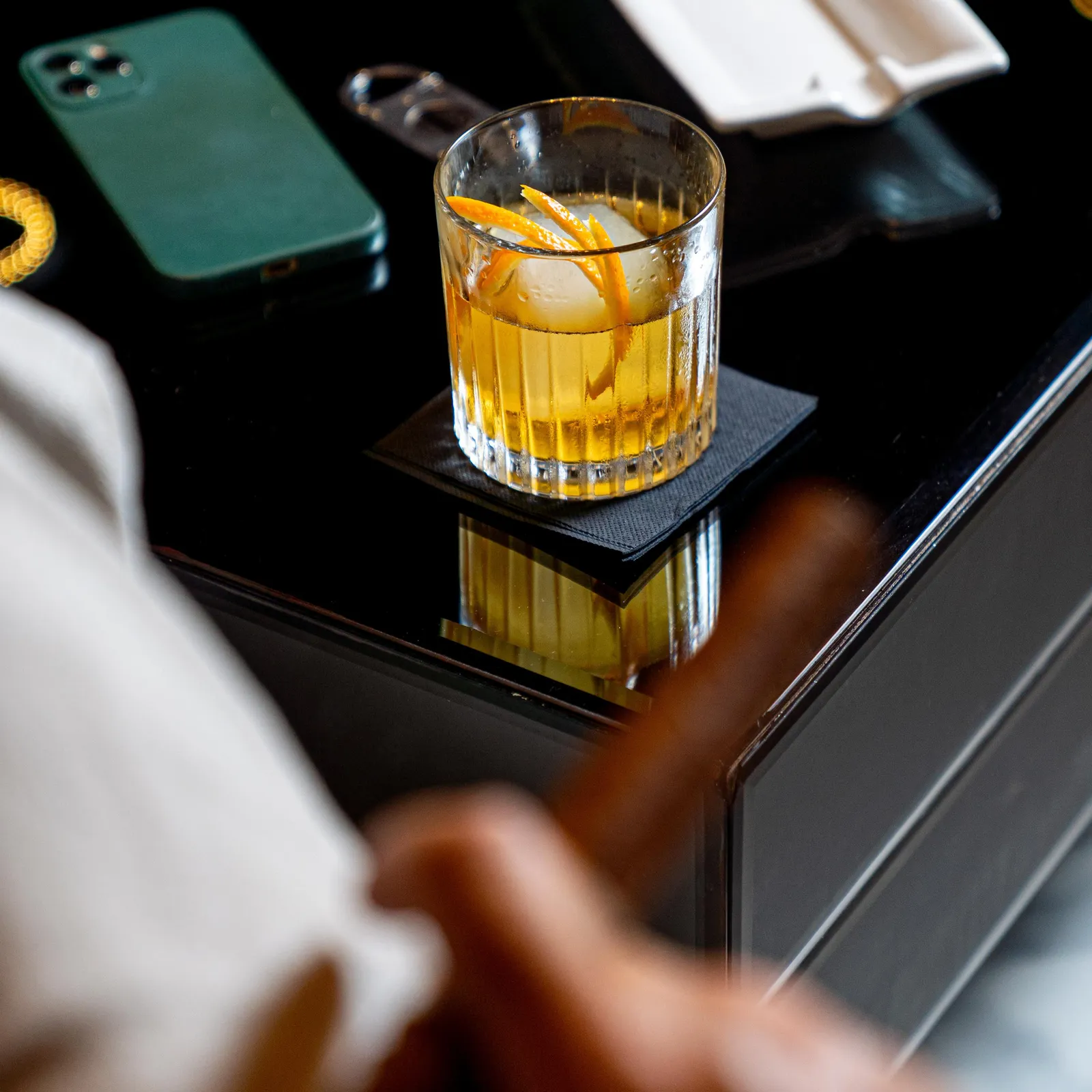 A glass containing a yellow liquid with ice cubes and an orange slice is placed on a black table, next to a green smartphone.