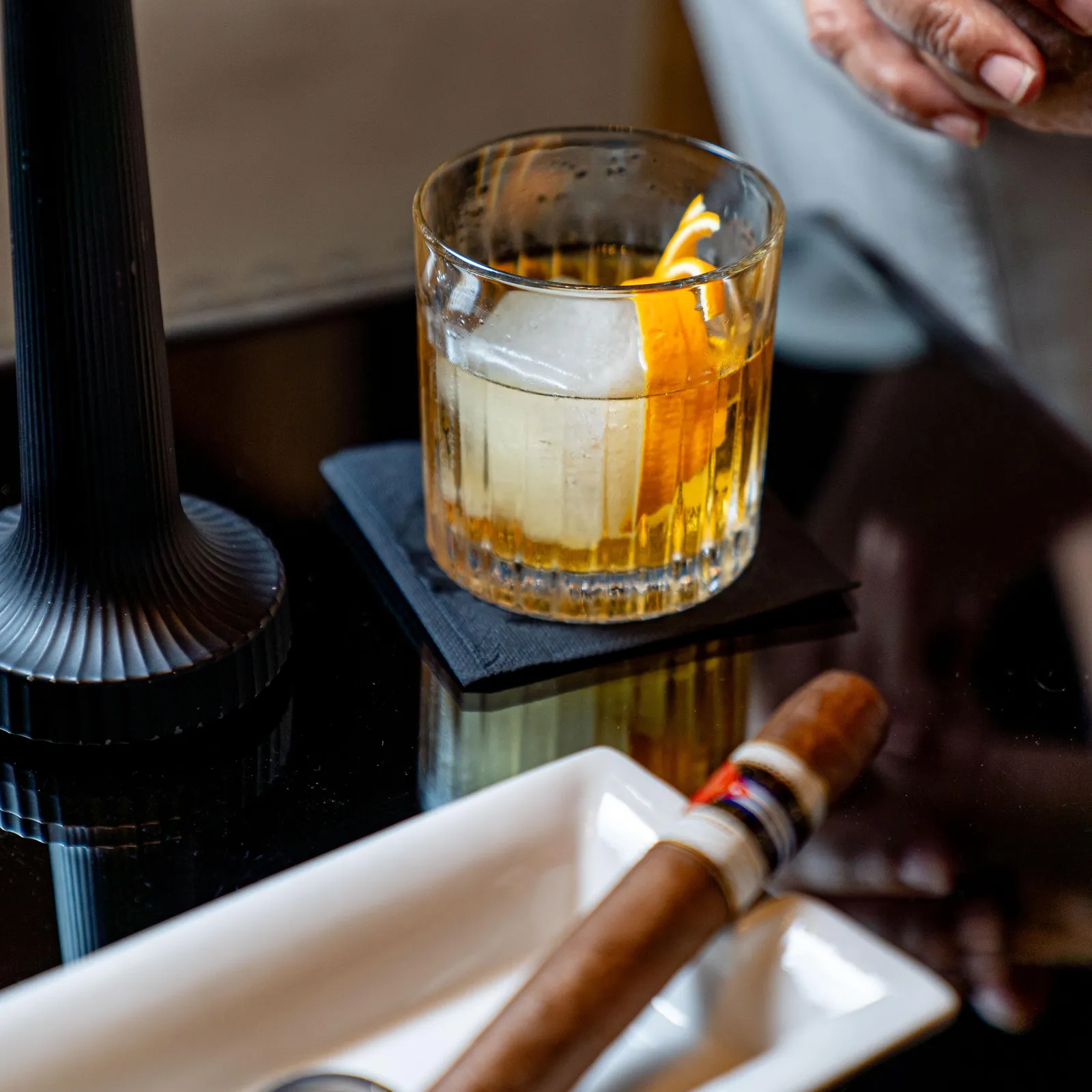 A glass containing an amber liquid with ice cubes and a slice of citrus, a cigar in a case, and hands resting on a table.