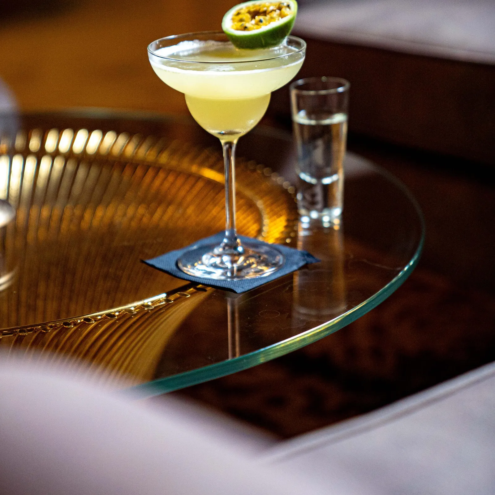 A cocktail glass containing a clear drink with a fruit slice on the rim, accompanied by a small glass of water, on a glass table.