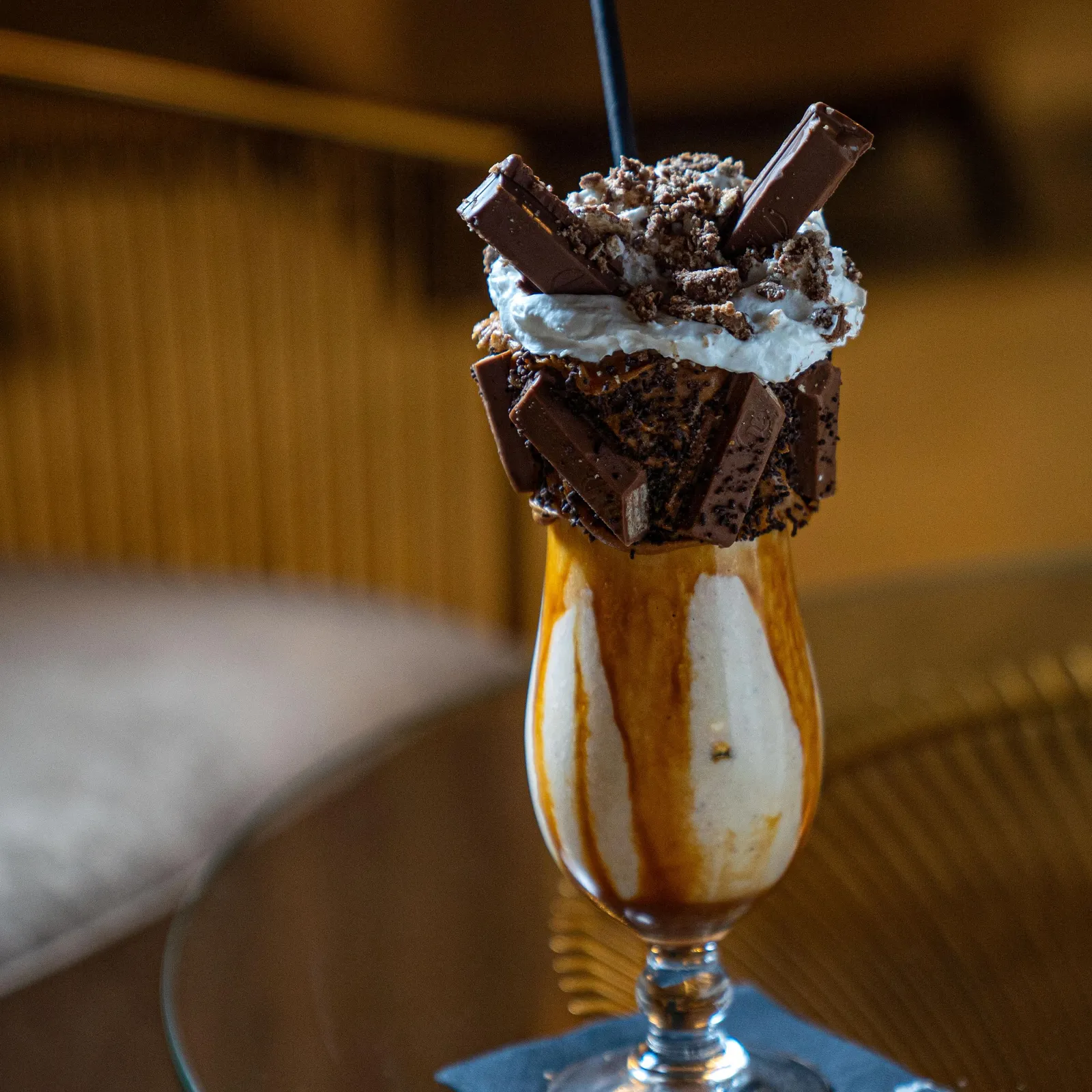 A transparent glass contains a drink with a garnish of whipped cream, biscuit pieces, and chocolate, placed on a metal saucer.