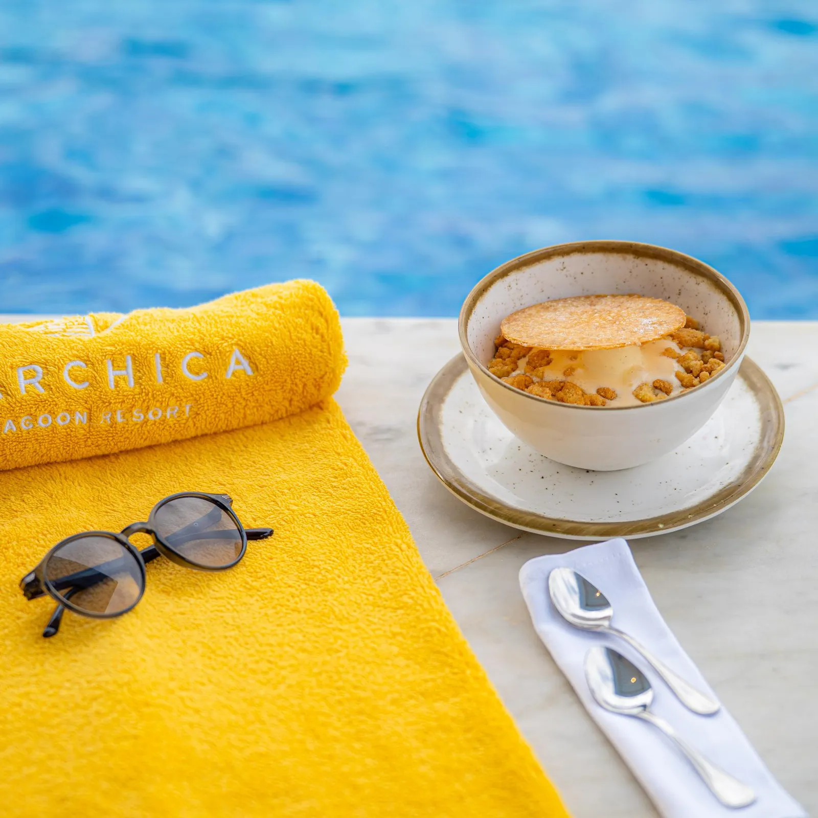 A glass of dessert with a crunchy layer, a yellow napkin, sunglasses, and a spoon on a table near a pool.
