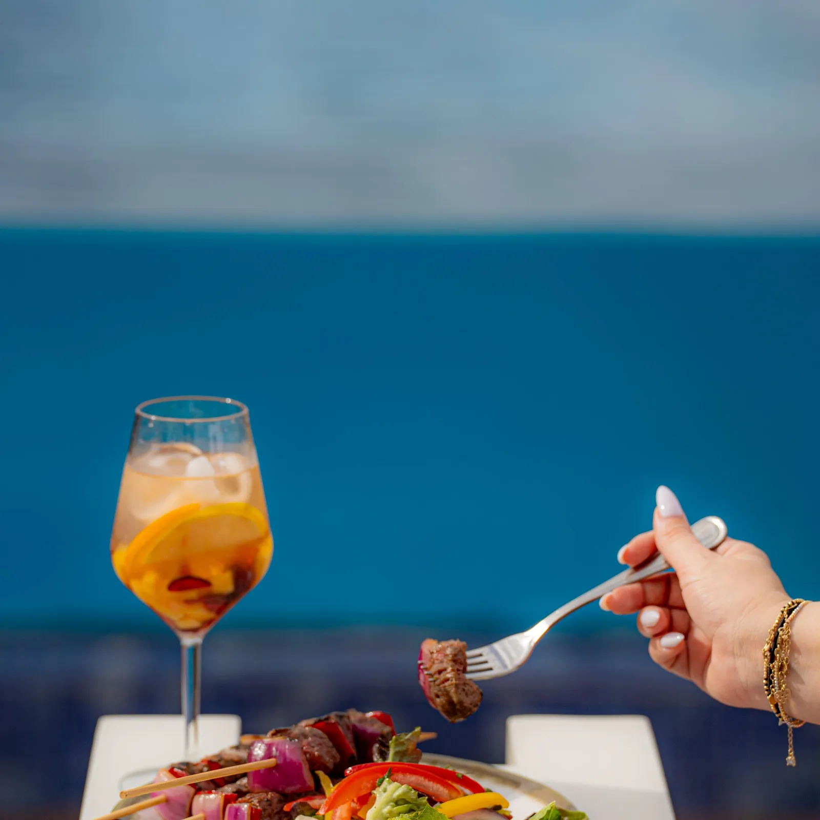A hand holds a fork with a piece of meat above a plate of skewers and salad, with an orange drink in the background in front of a blue sky.