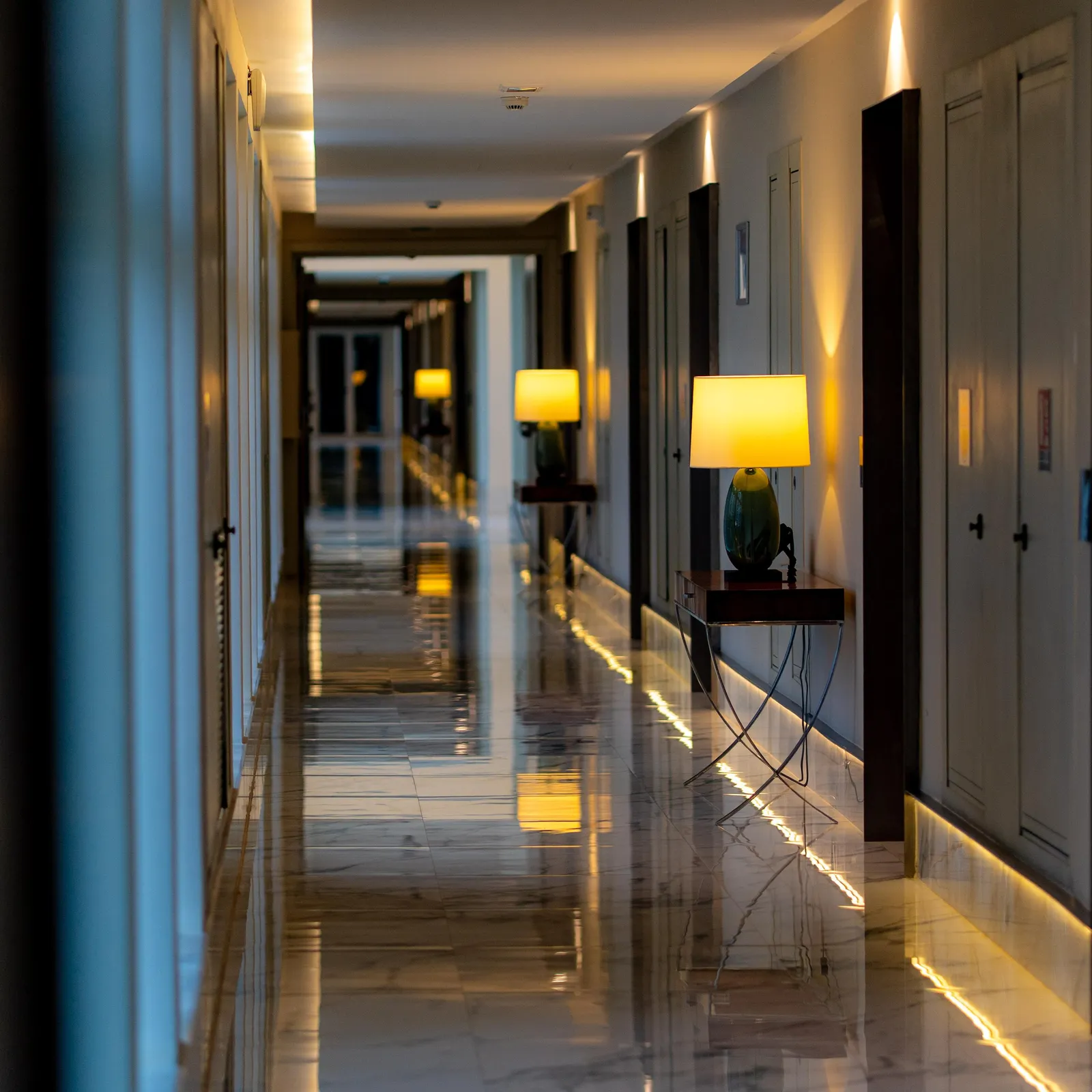 An interior hallway lit by lamps on low tables, reflected by a shiny floor