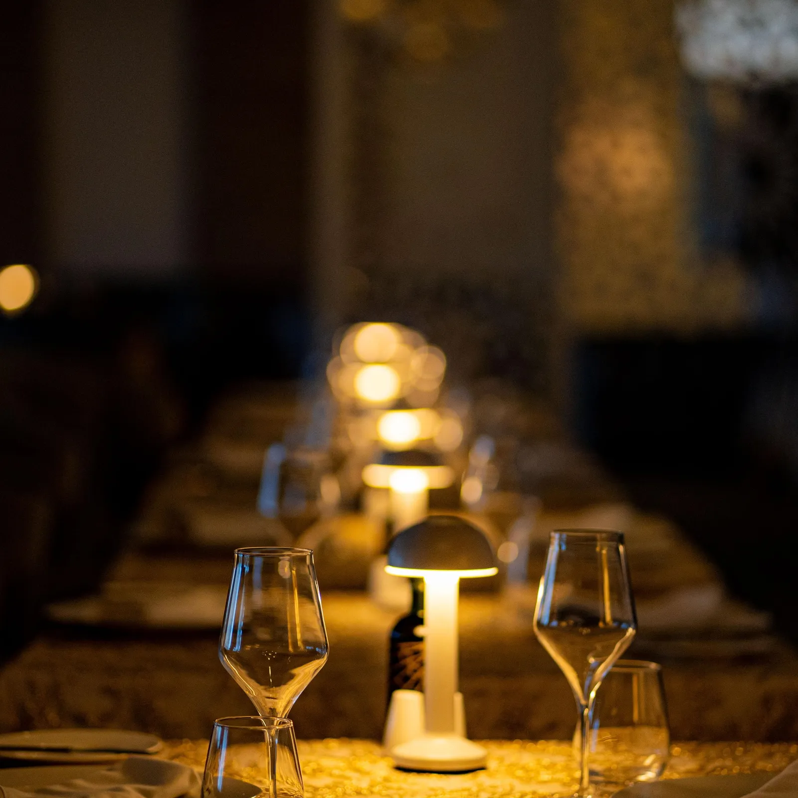 Table set with wine glasses, tablecloth, and candles, lit by lamps in the background