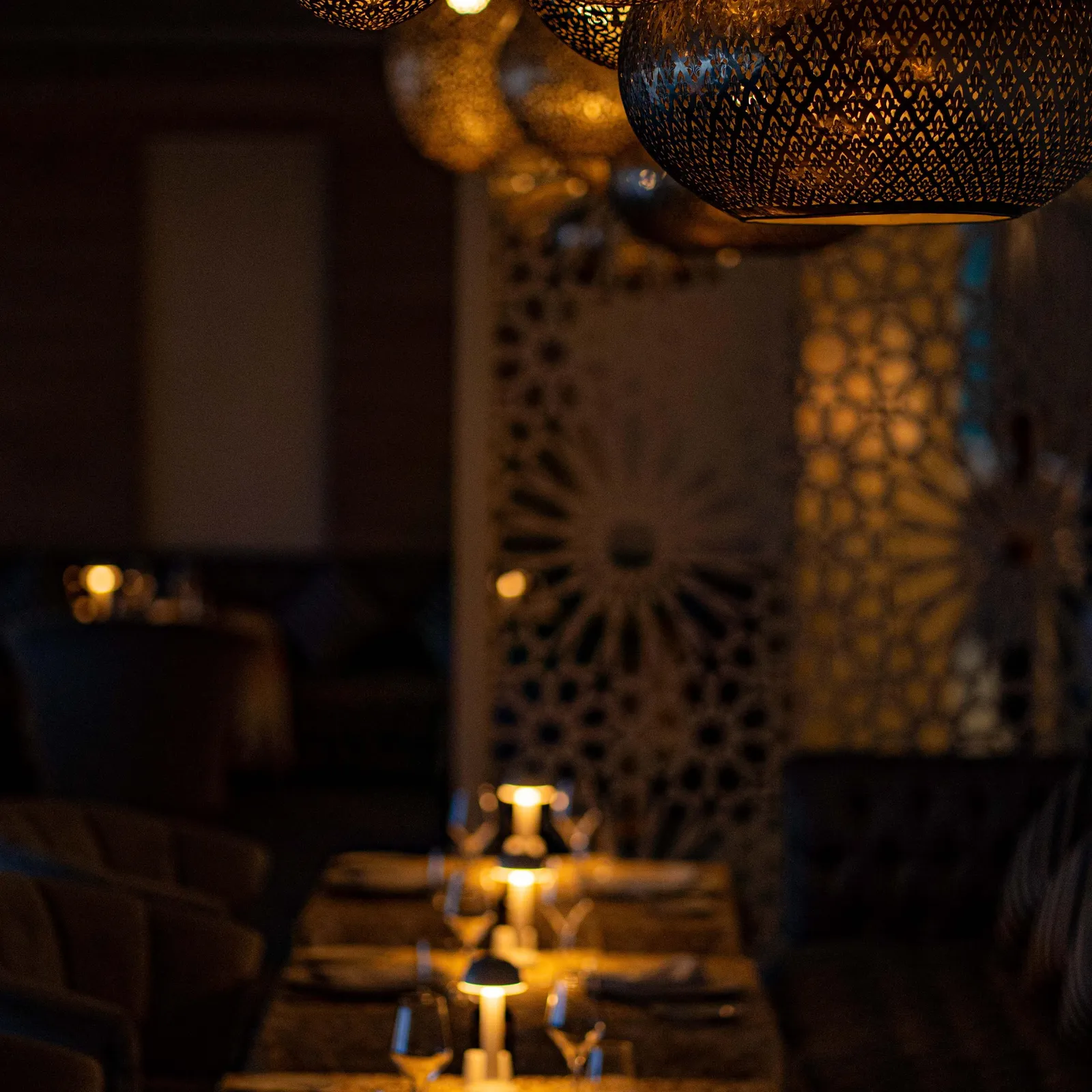A table set with glasses, candles, and cutlery under light fixtures in a dimly lit restaurant.