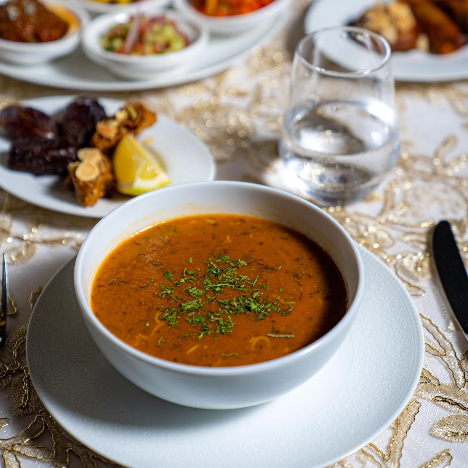 A bowl of orange soup with fresh herbs placed on a white plate, surrounded by several small dishes containing various foods, all on a table covered with a golden tablecloth.