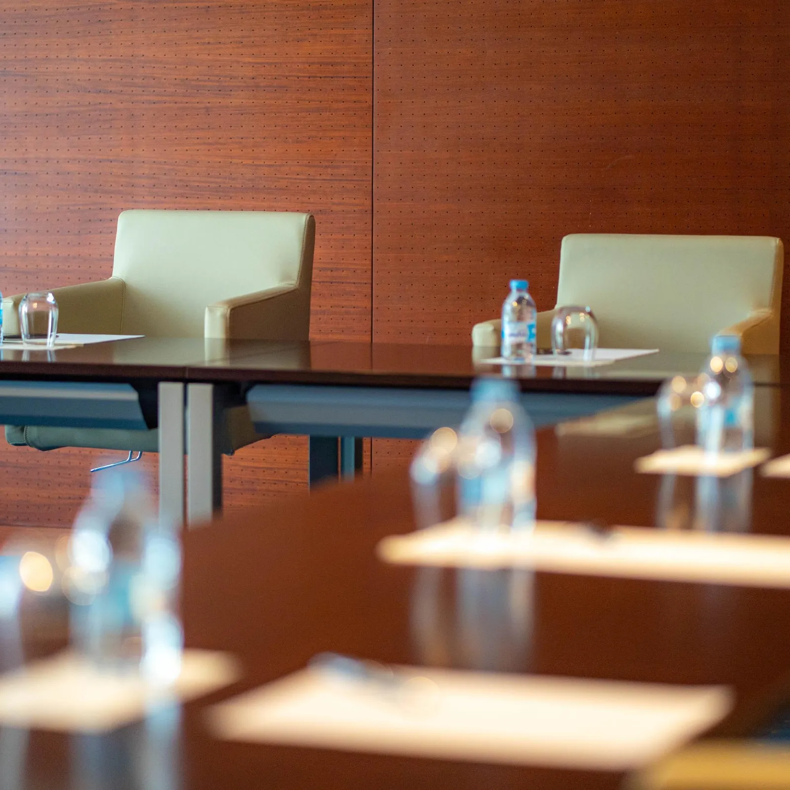 A row of conference tables with water bottles and glasses in front of empty chairs in a room with wooden walls