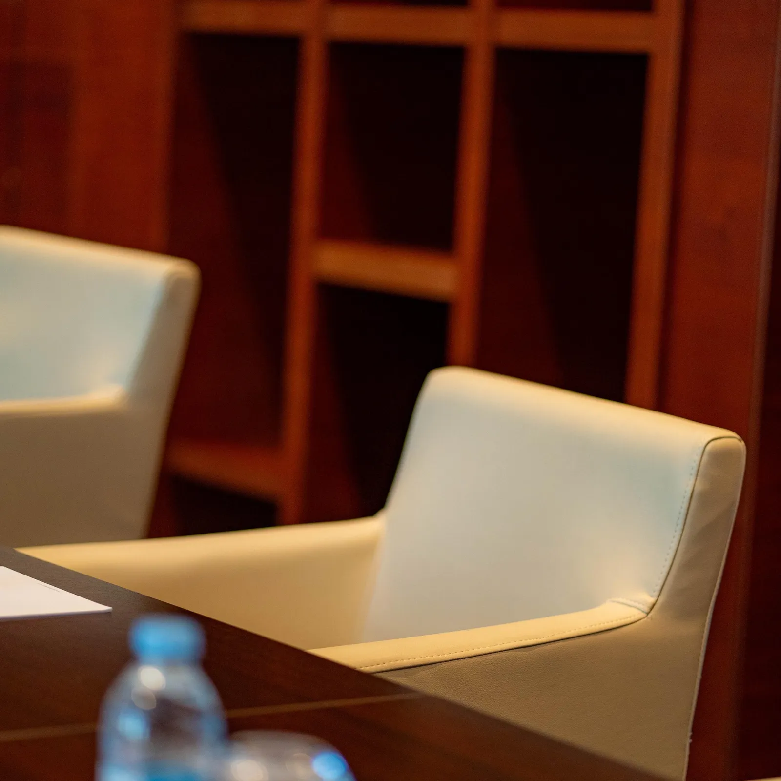 A bottle of water and a pair of glasses are placed on a table in front of a beige leather chair in a room with dark wooden panels.
