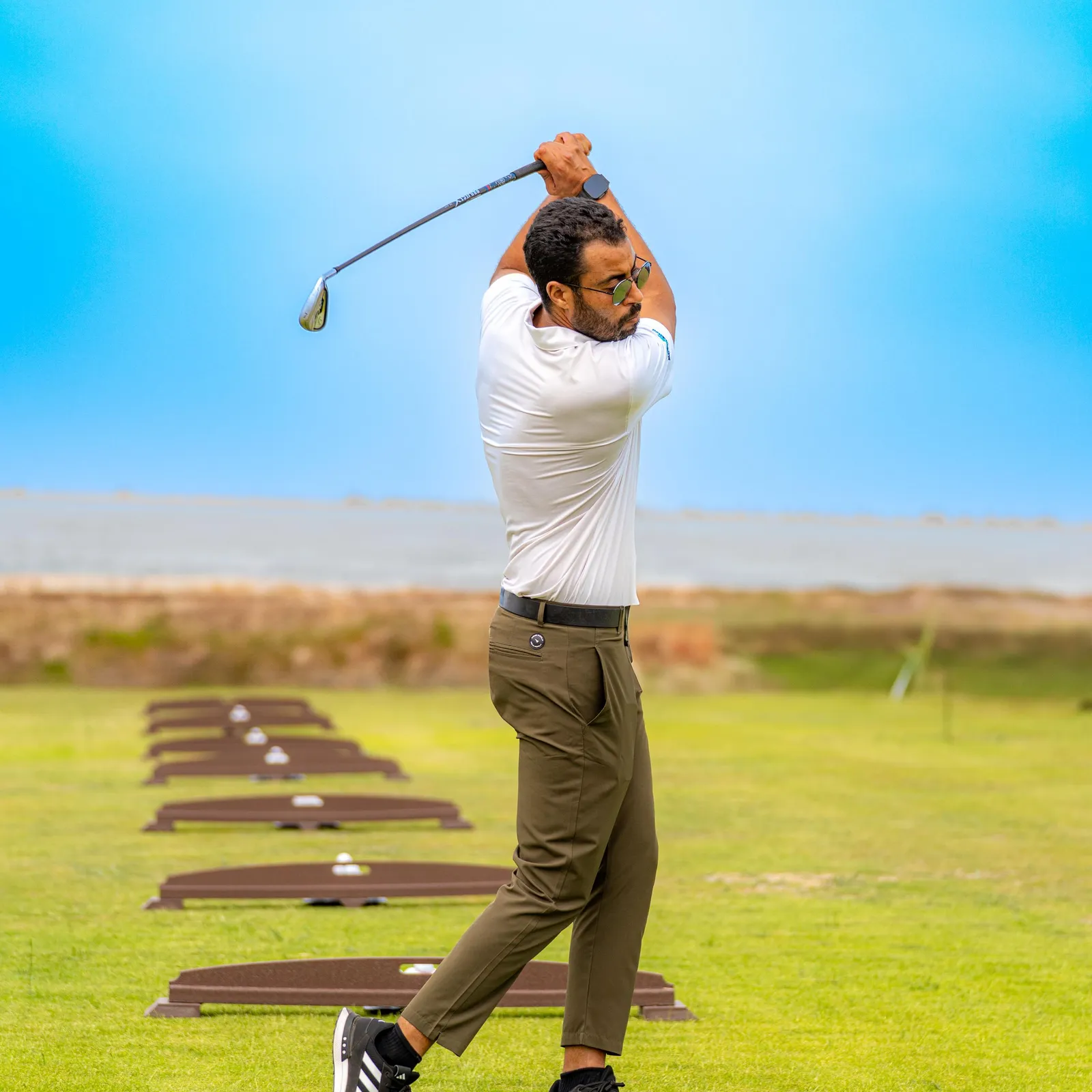 A man in a white polo shirt and green pants hits golf balls on an outdoor practice range.