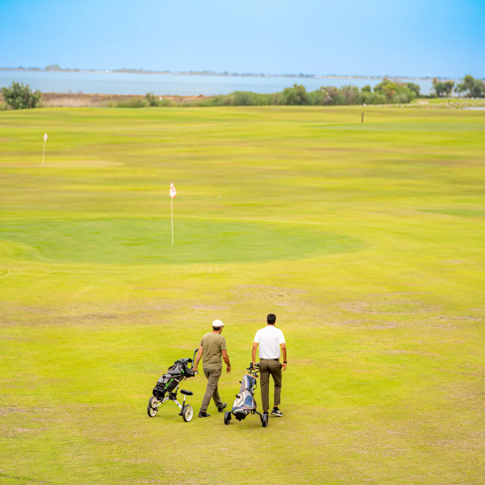 Two people with golf carts walk on a golf green with a flag visible in the distance and a body of water in the background.